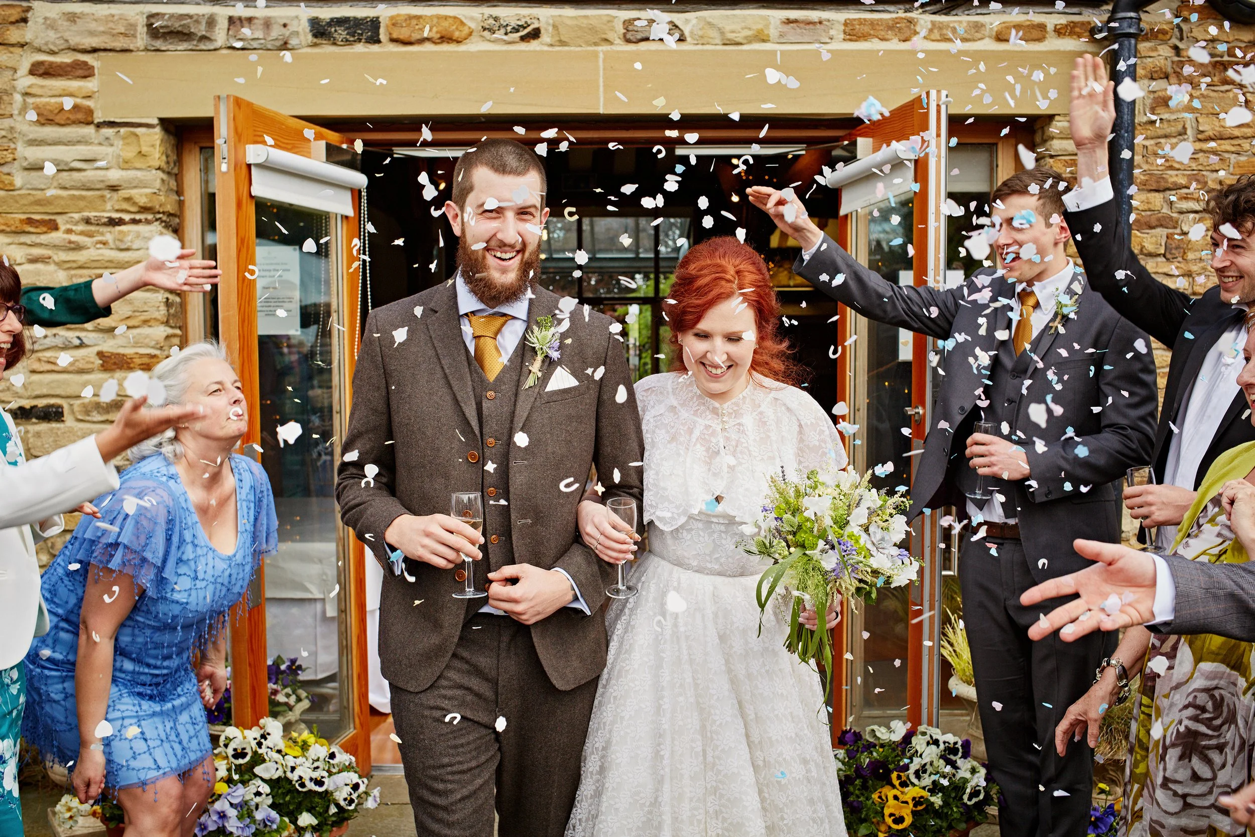 A bride and groom celebrating their wedding surrounded by friends and family as confetti falls, standing outside a building with a stone wall and glass door.