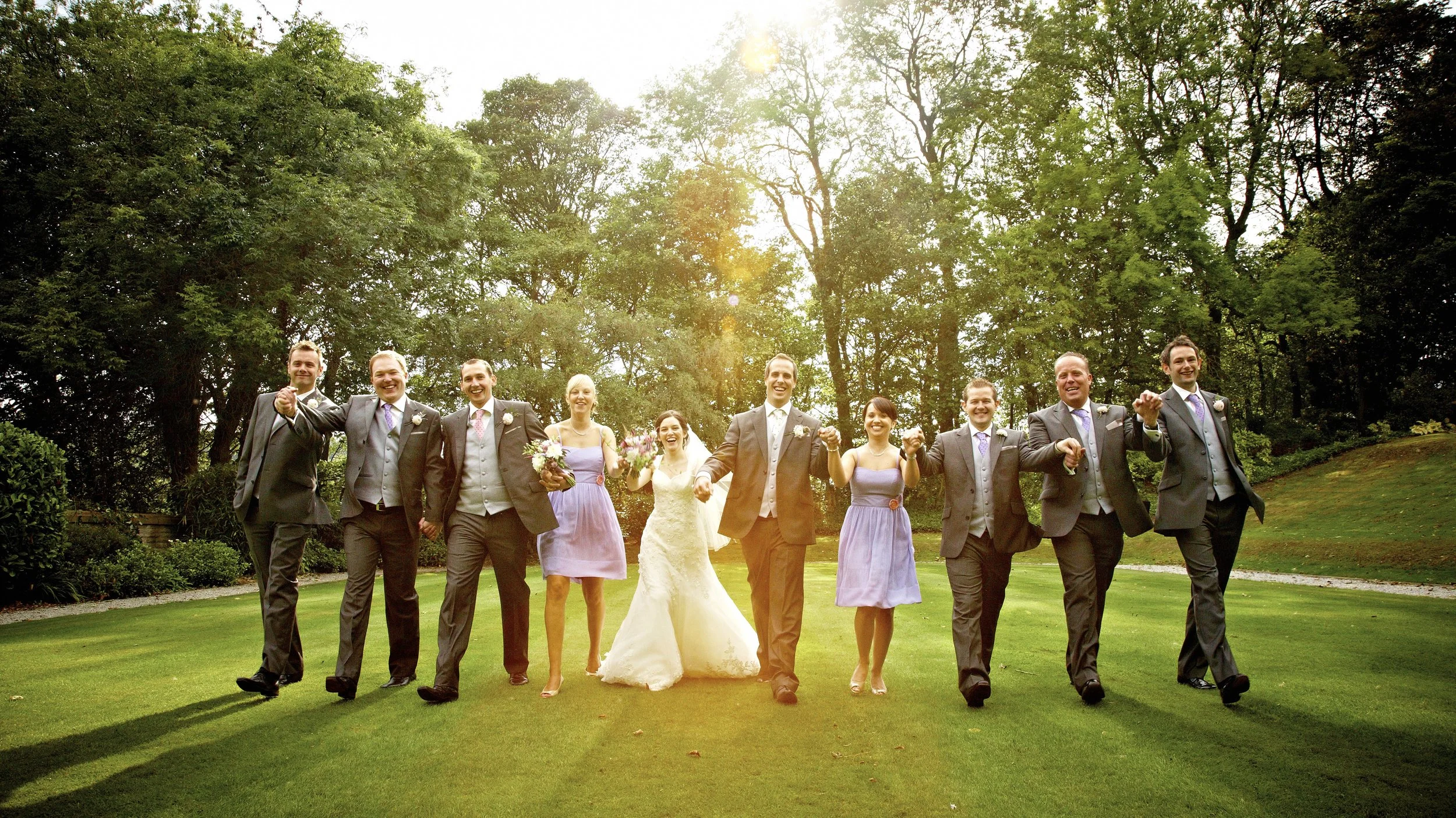A group of wedding guests and a bride and groom walking outdoors on a sunny day, holding hands and smiling.