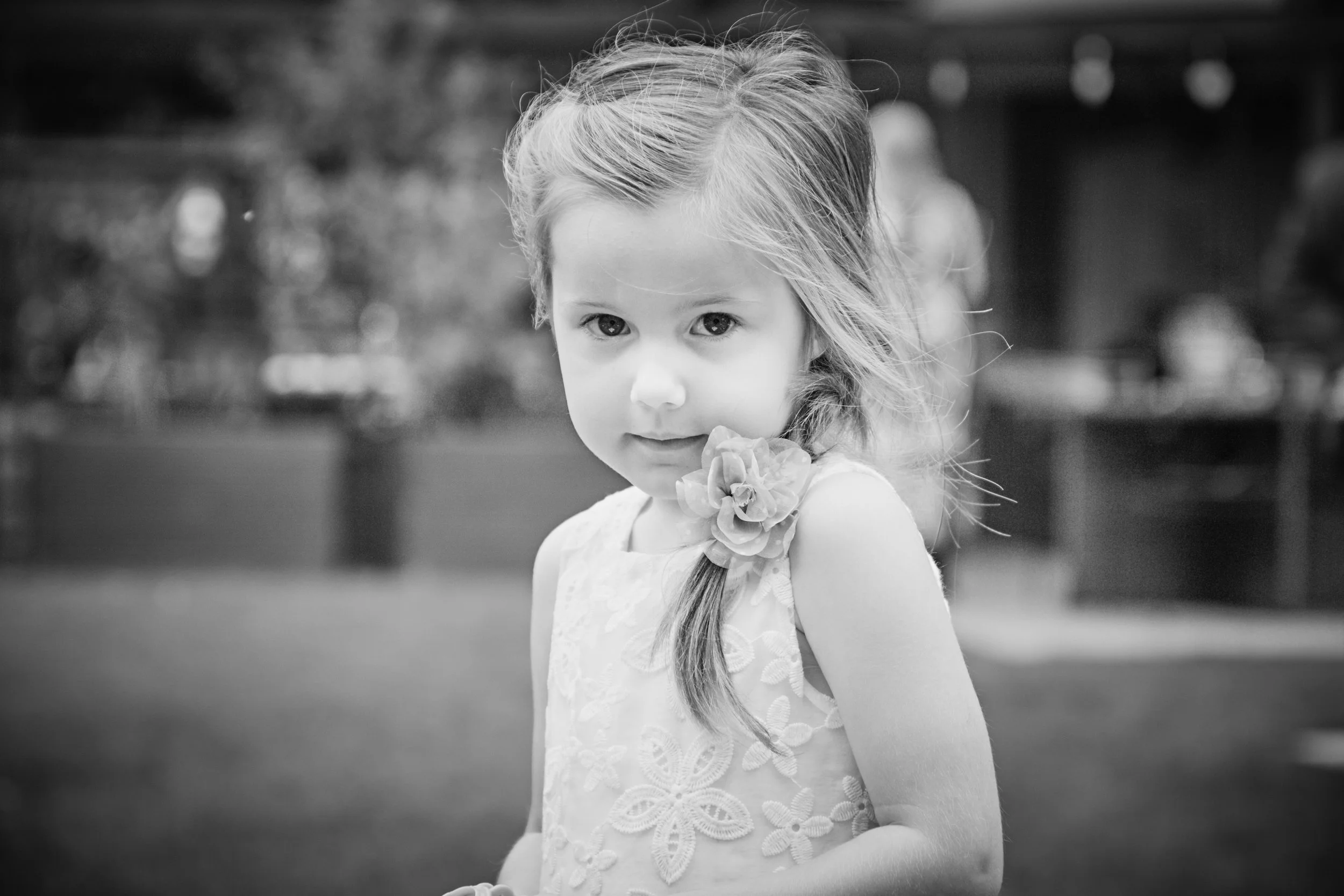Black and white photo of a young girl with long hair wearing a dress with floral embroidery, holding a flower near her face, outdoors.
