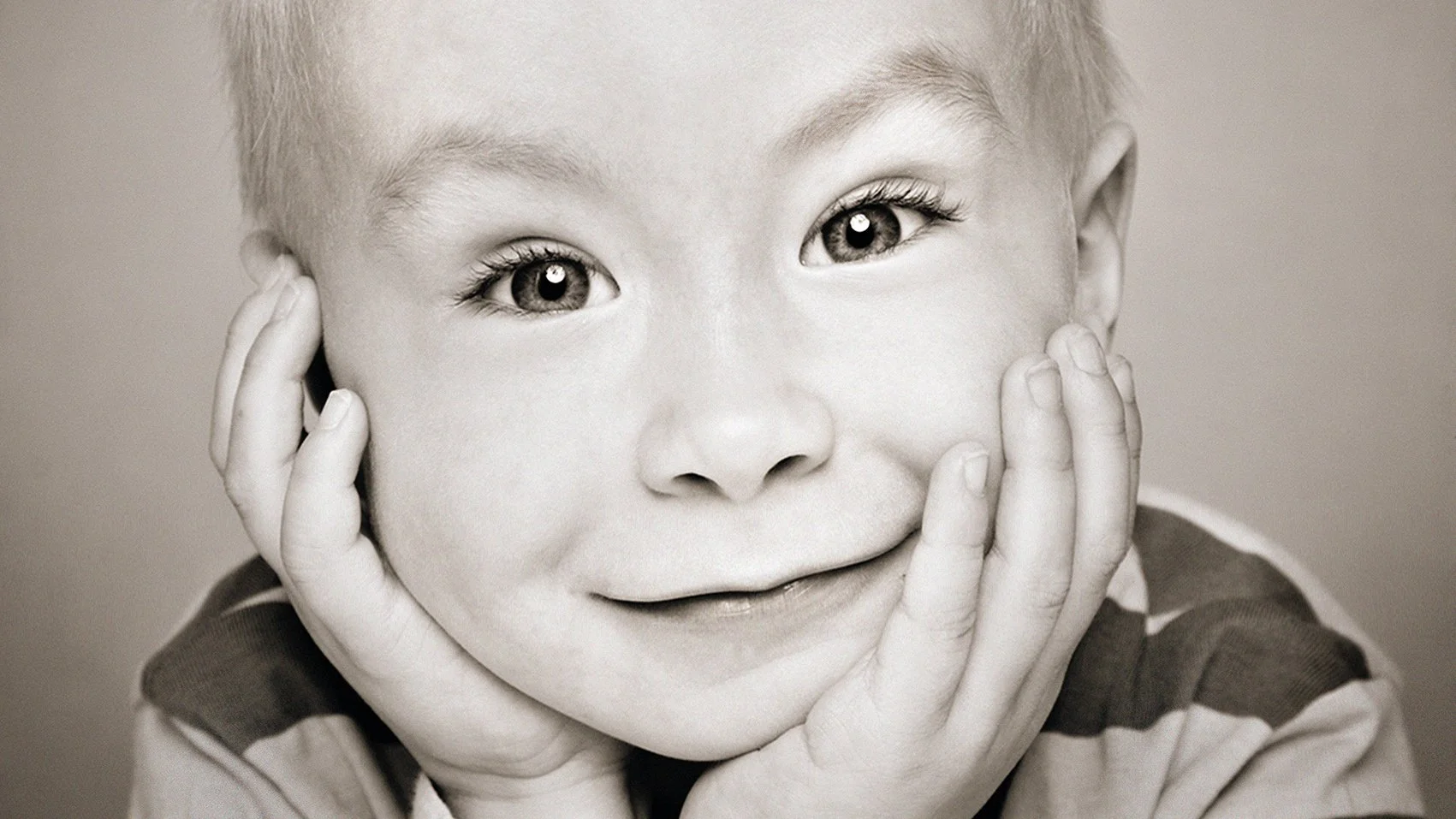 A close-up black and white photo of a smiling child with hands on cheeks, showing expressive eyes and a joyful expression.