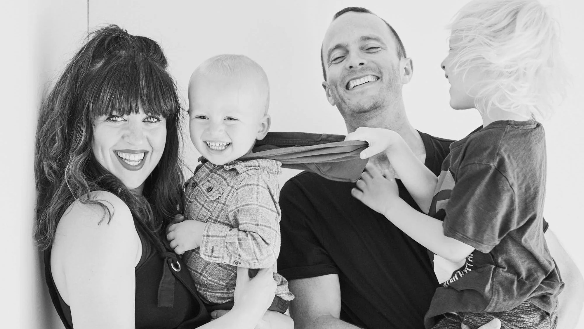 A happy family of four, including a woman, a man, and two young children, playing together and smiling in a black and white photo.