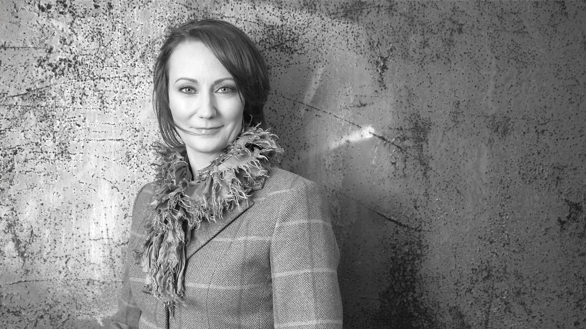 A woman with short hair smiling, wearing a patterned jacket and a ruffled scarf, standing against a textured concrete wall.