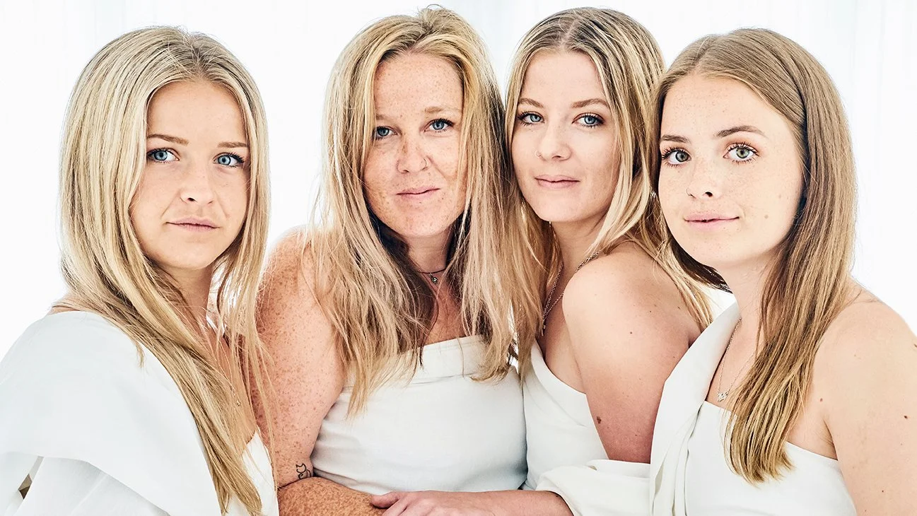 Four women with blonde hair and fair skin wearing white clothing standing close together against a white background.