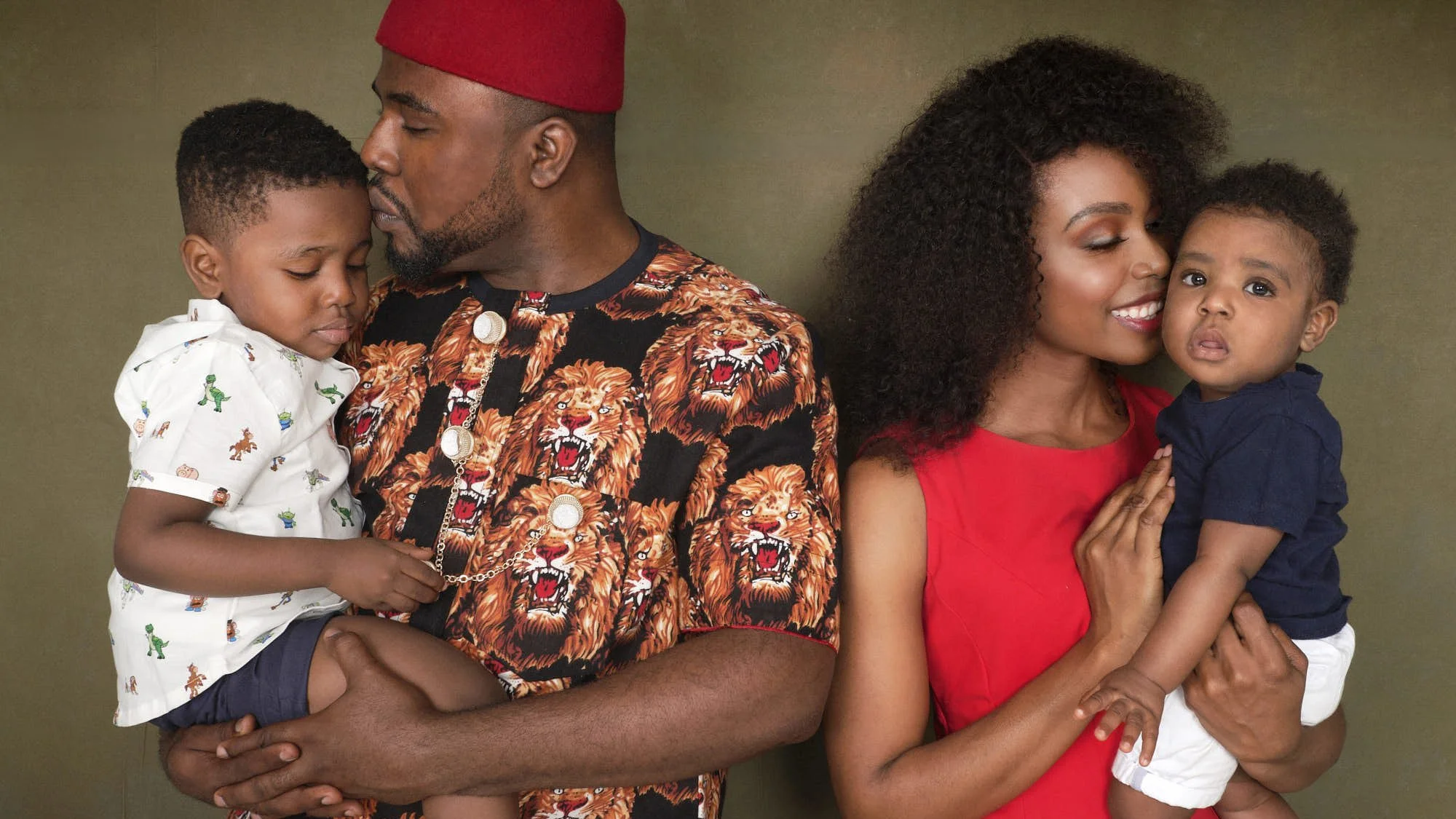 A smiling woman with curly hair holding a young boy. A man with a beard in a lion-themed shirt is holding a young boy who is resting his head on his shoulder, with both boys having curly hair.