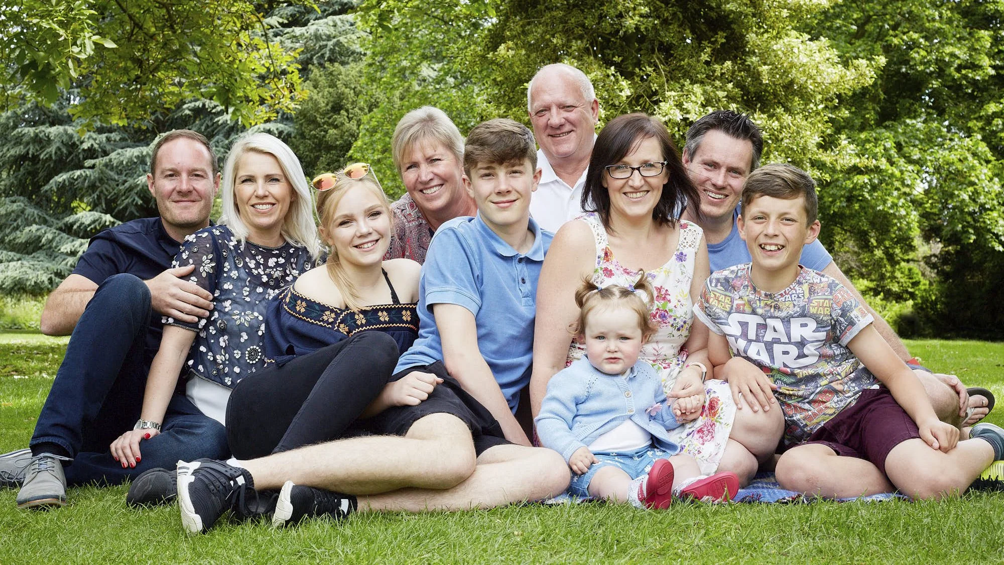 Group of smiling people, including children and adults, sitting on grass outdoors among trees in a park.