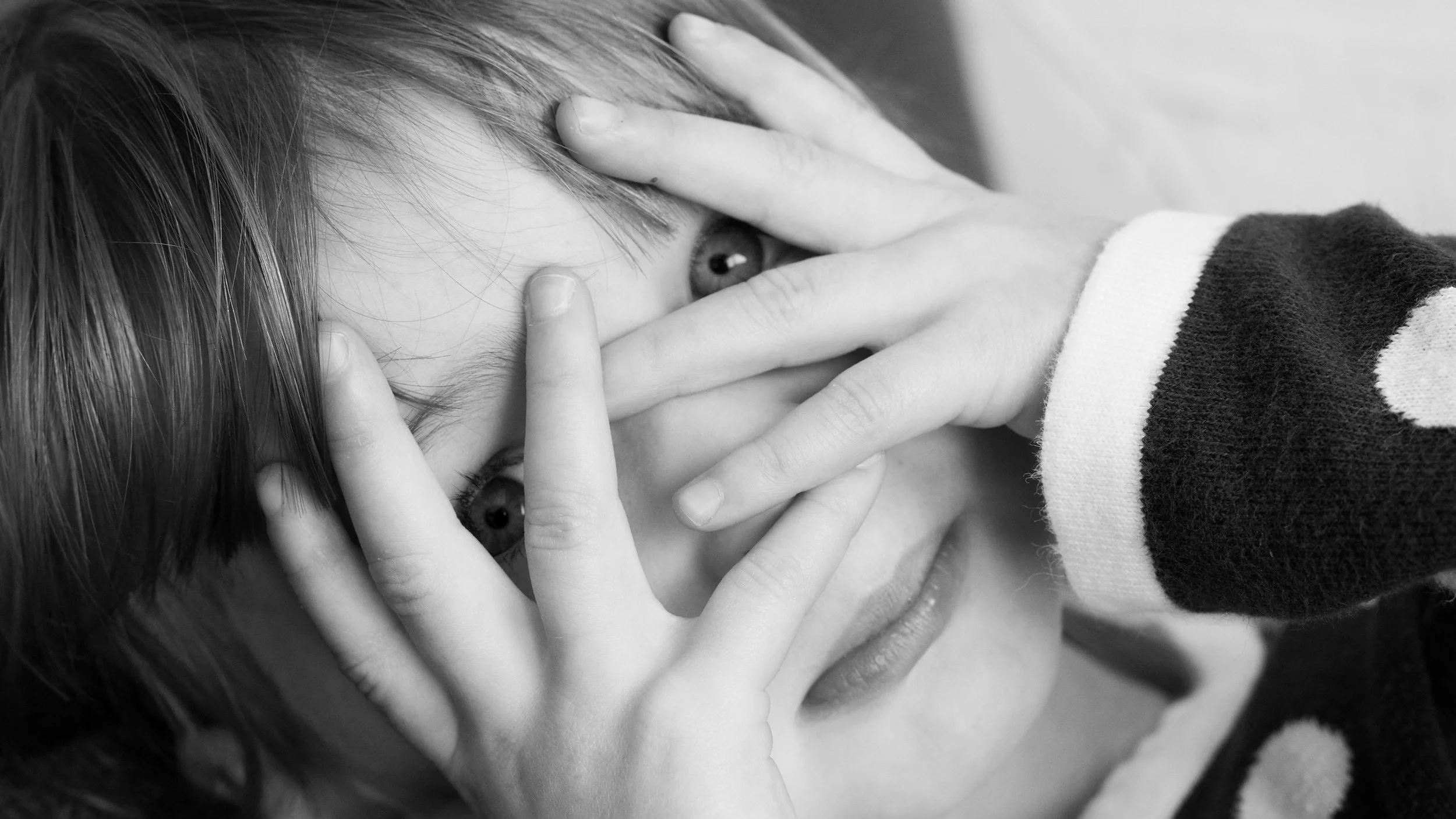 Black and white photo of a person holding their face with both hands, partially covering one eye, with long hair and a striped sleeve.