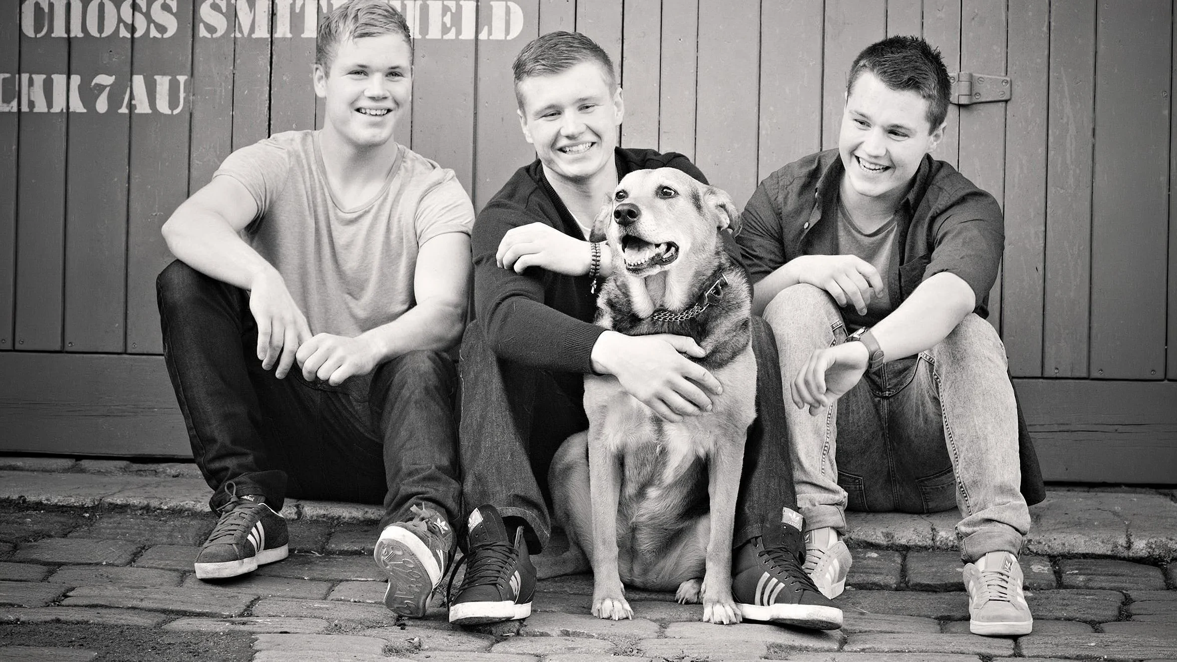 Four young men sitting on a curb with a dog in front of a wooden wall, smiling and looking at the camera, in black and white.