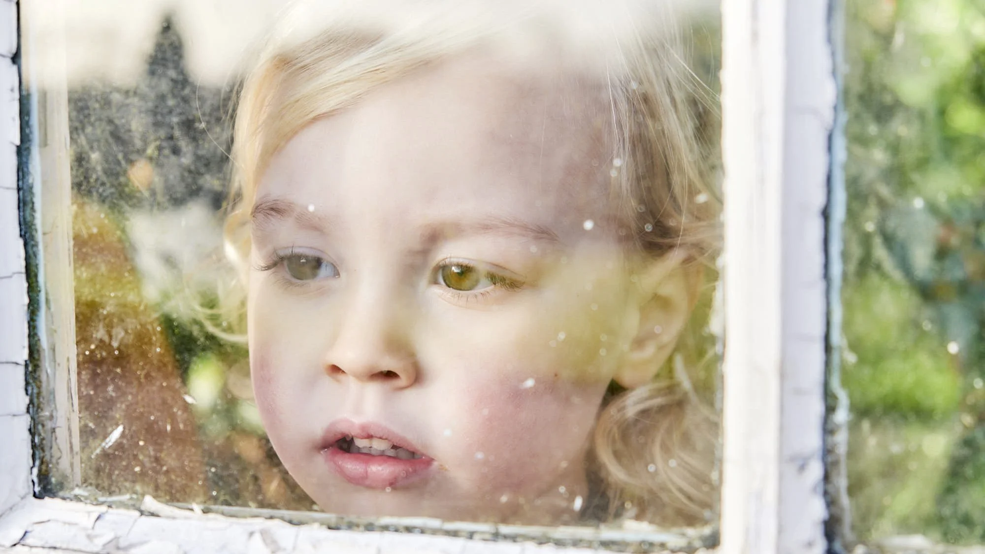 A young girl with blonde hair looking through a window with peeling paint, with green trees visible outside.