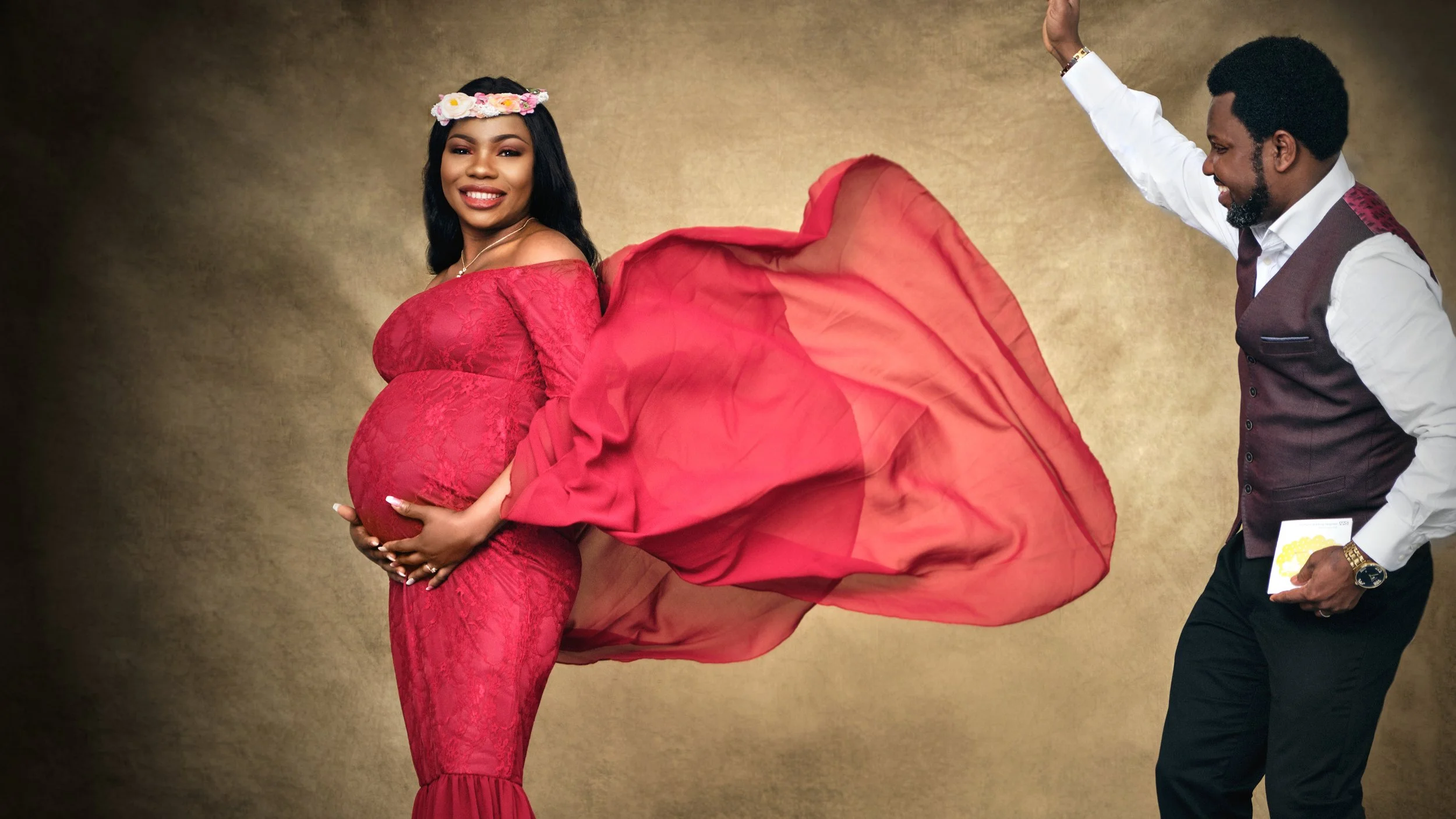 A pregnant woman in a red dress with lace details holds her belly and smiles, while a man in a white shirt and vest playfully holds a large piece of red fabric or dress behind her, against a gold background.
