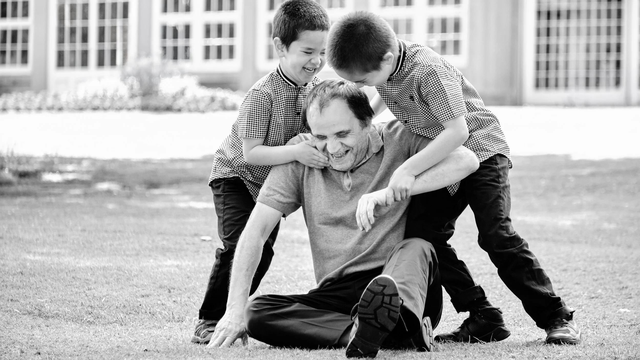 Two young boys playfully holding down an adult man on a grassy field, all smiling and laughing while engaging in a fun outdoor activity.