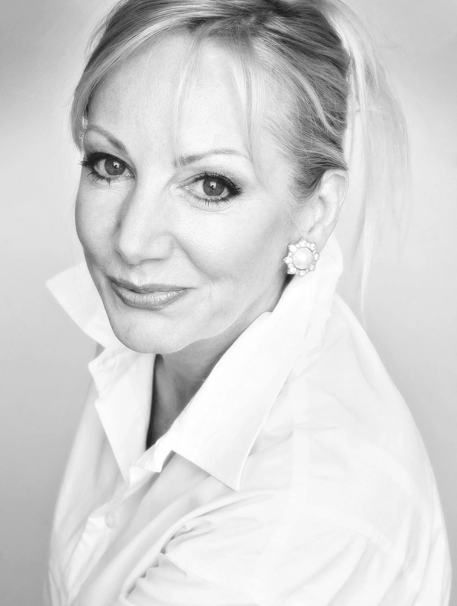 Black-and-white portrait of a woman wearing a white shirt and large pearl earrings, with her hair styled back, smiling softly.