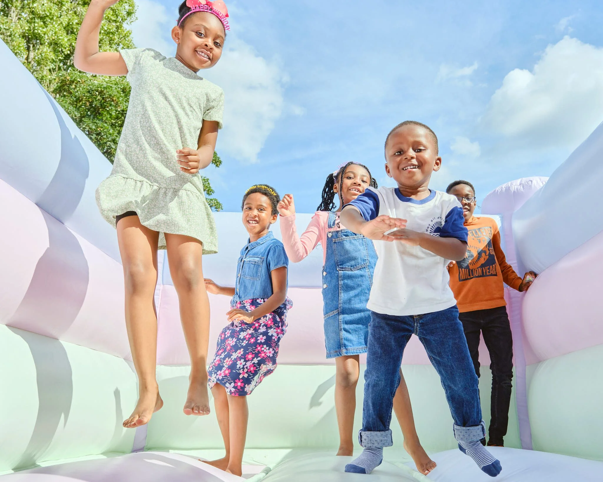 Group of children playing indoors, standing on a large inflatable bounce house with blue sky and green trees outside.