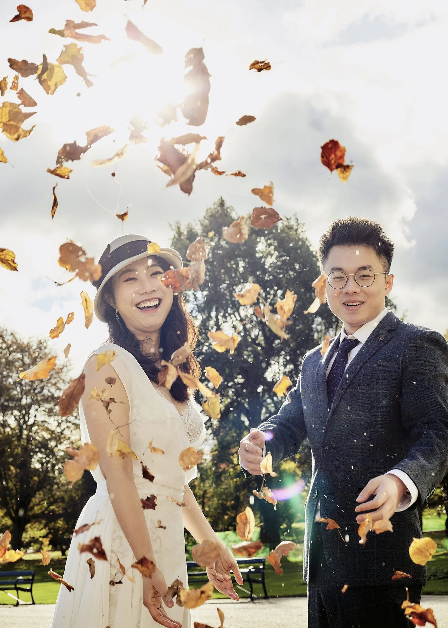 A woman in a white dress and hat smiling and a man in a dark suit and glasses smiling, holding hands, with autumn leaves falling around them outdoors on a sunny day.