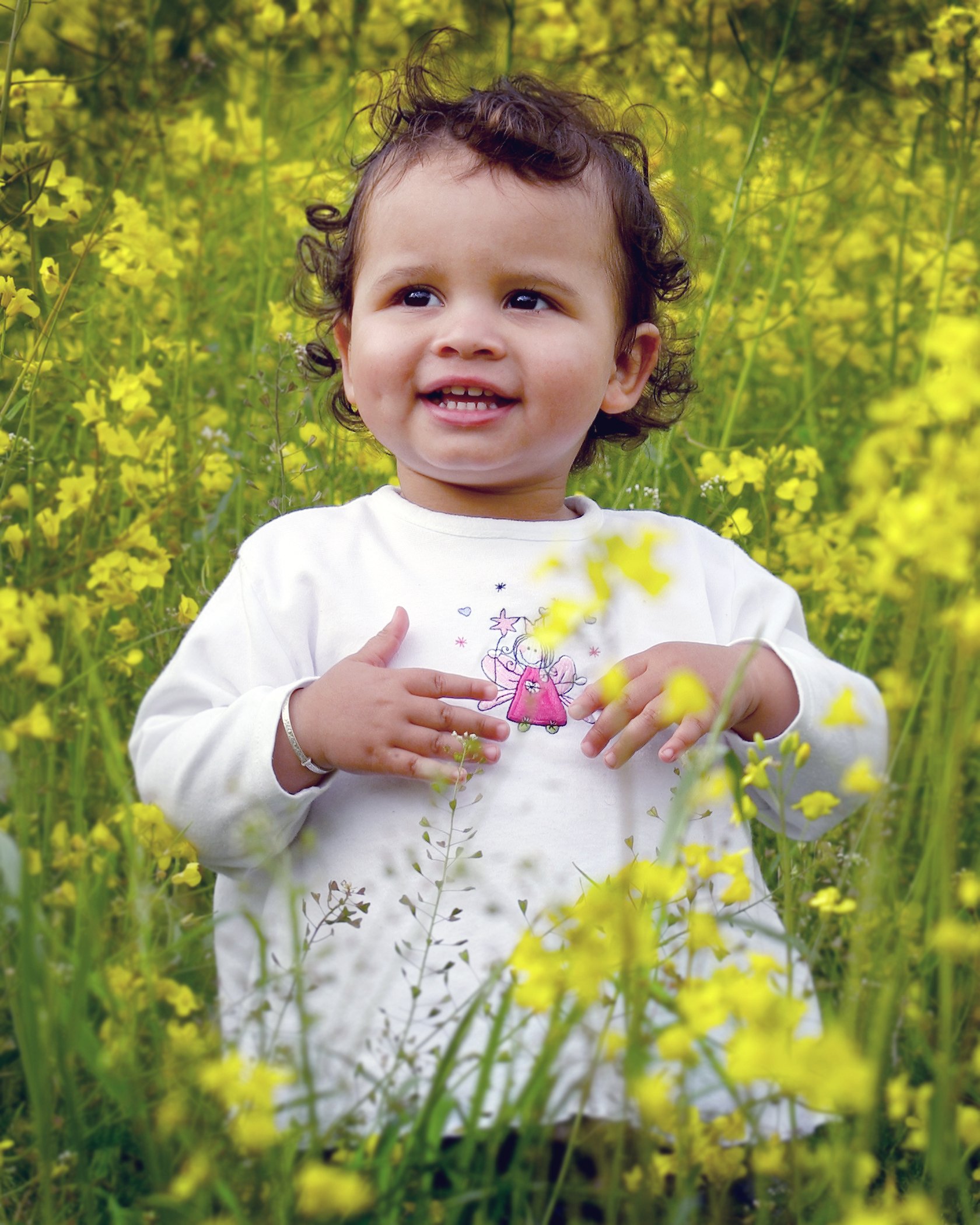 A young child with curly hair and a big smile, wearing a white shirt with a cartoon character, standing amidst yellow flowers in a green field.