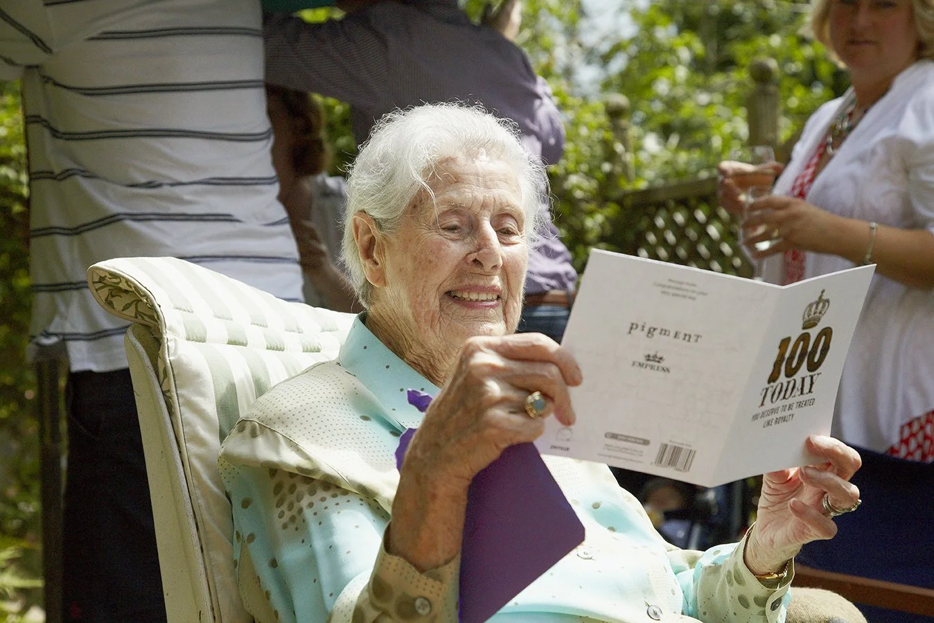 A senior woman with white hair sitting in a garden chair, reading a birthday card that says '100 Today' and smiling, with people in the background holding drinks.