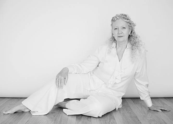 Black and white photograph of a woman with curly hair sitting on the floor against a plain background, wearing a white blouse and pants.