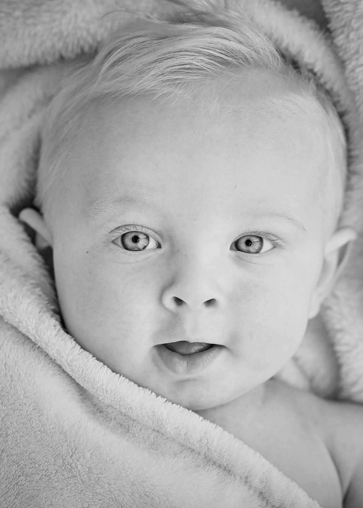 Close-up black and white photo of a baby's face, showing their wide open eyes, a small nose, and slightly open mouth, wrapped in a soft blanket.