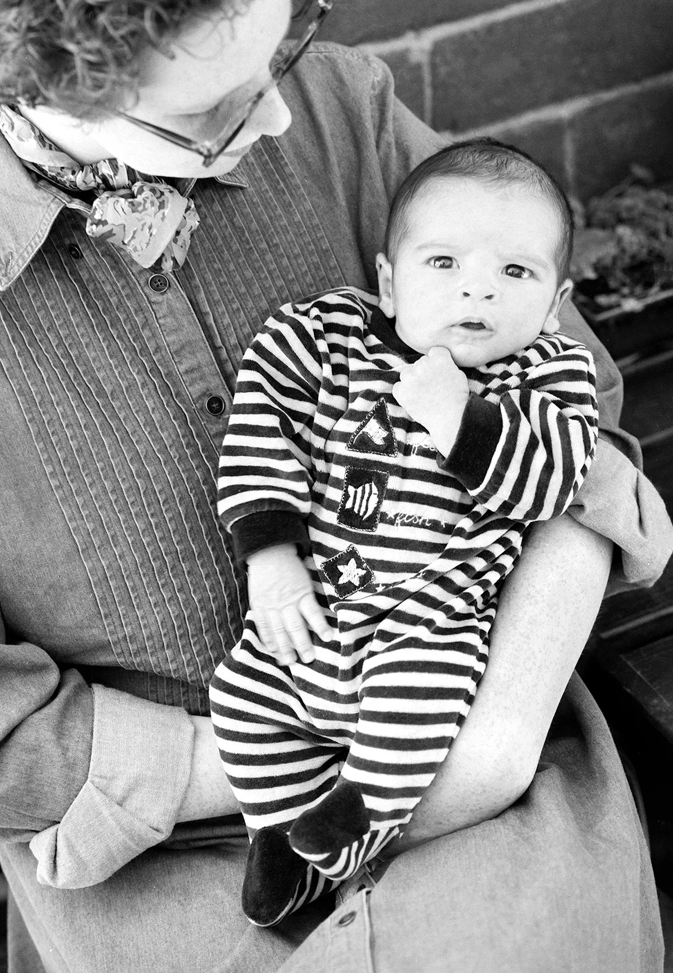 A baby wearing a striped onesie sitting on an adult's lap, holding their chin, with an adult wearing glasses and a button-up shirt nearby. The scene is in black and white.