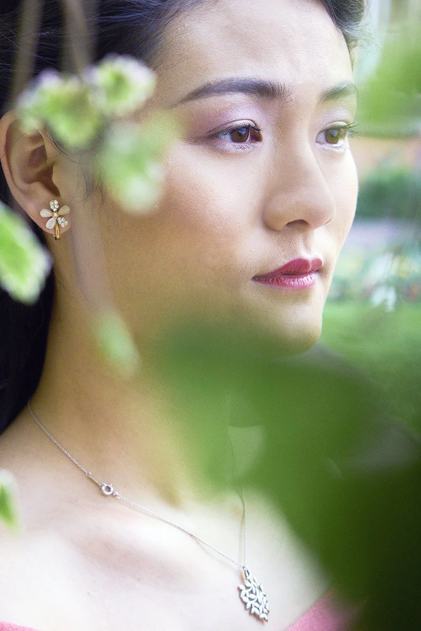 A young woman with makeup and jewelry, looking thoughtfully through green foliage, outdoors in a garden or park.