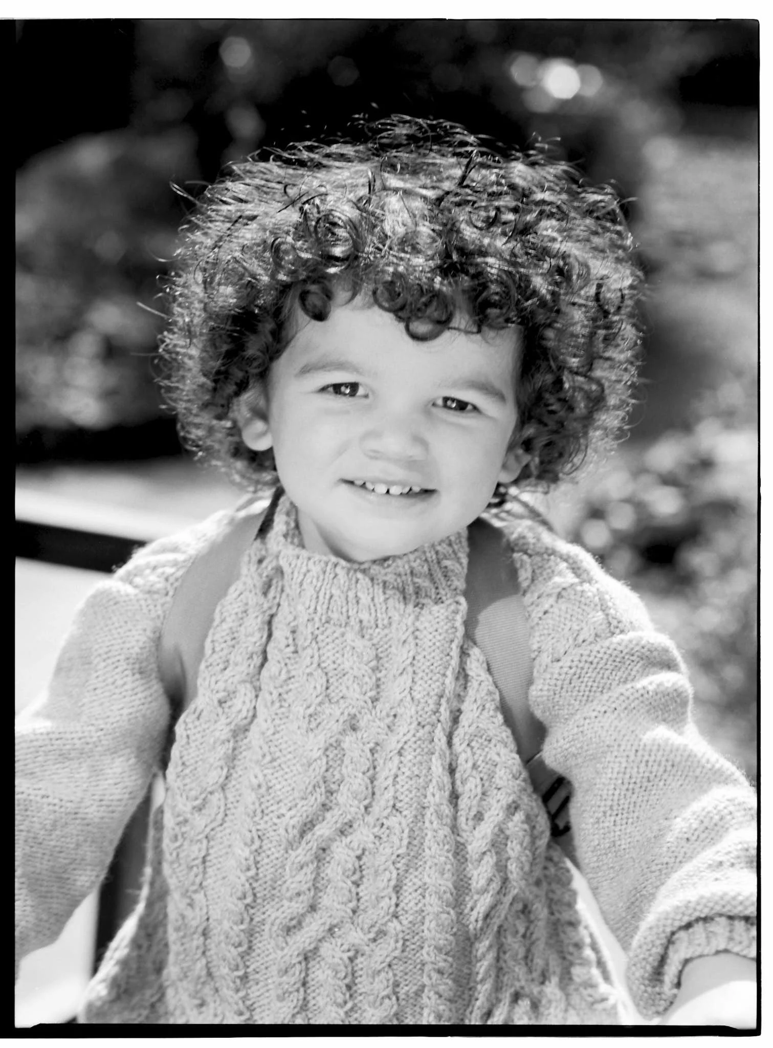 Black and white photo of a smiling young child with curly hair, wearing a knitted sweater, outdoors.