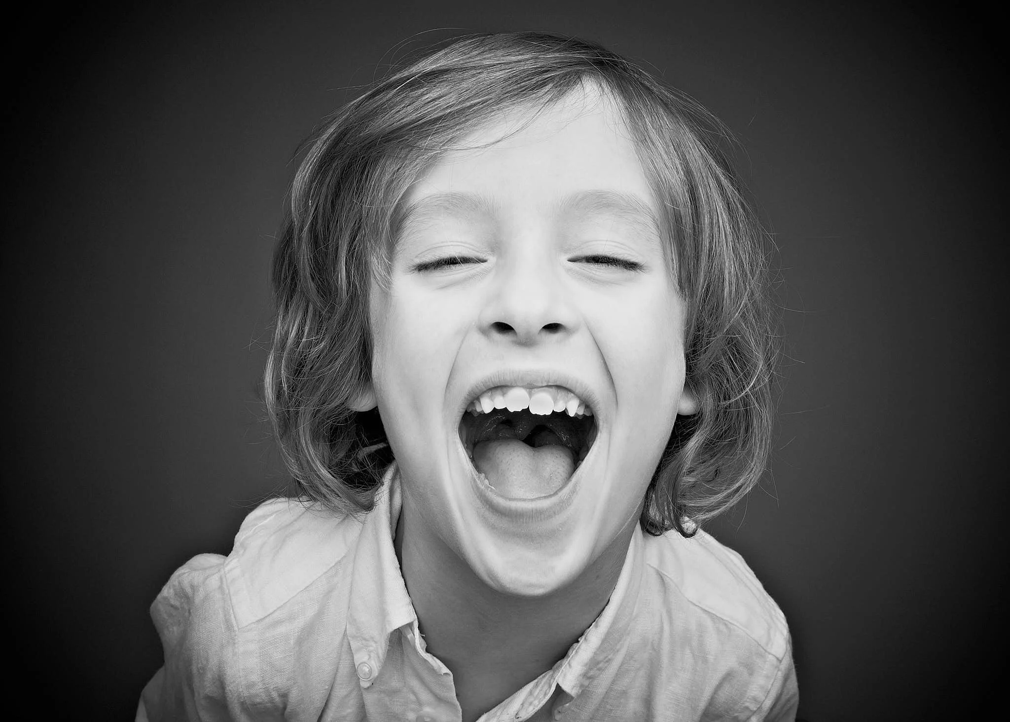 Black and white photo of a young child with an excited expression, mouth open, sticking out tongue, eyes squinting, wearing a collared shirt.