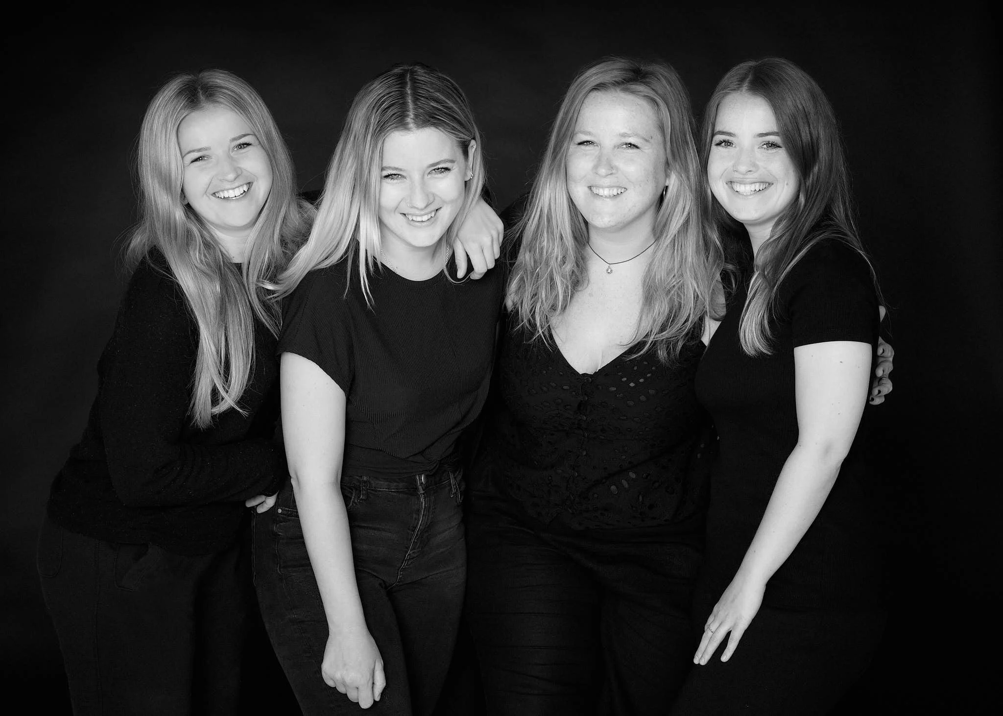 Black and white photo of four young women standing closely together, smiling, with their hands placed on the ground in front of them, against a dark background.