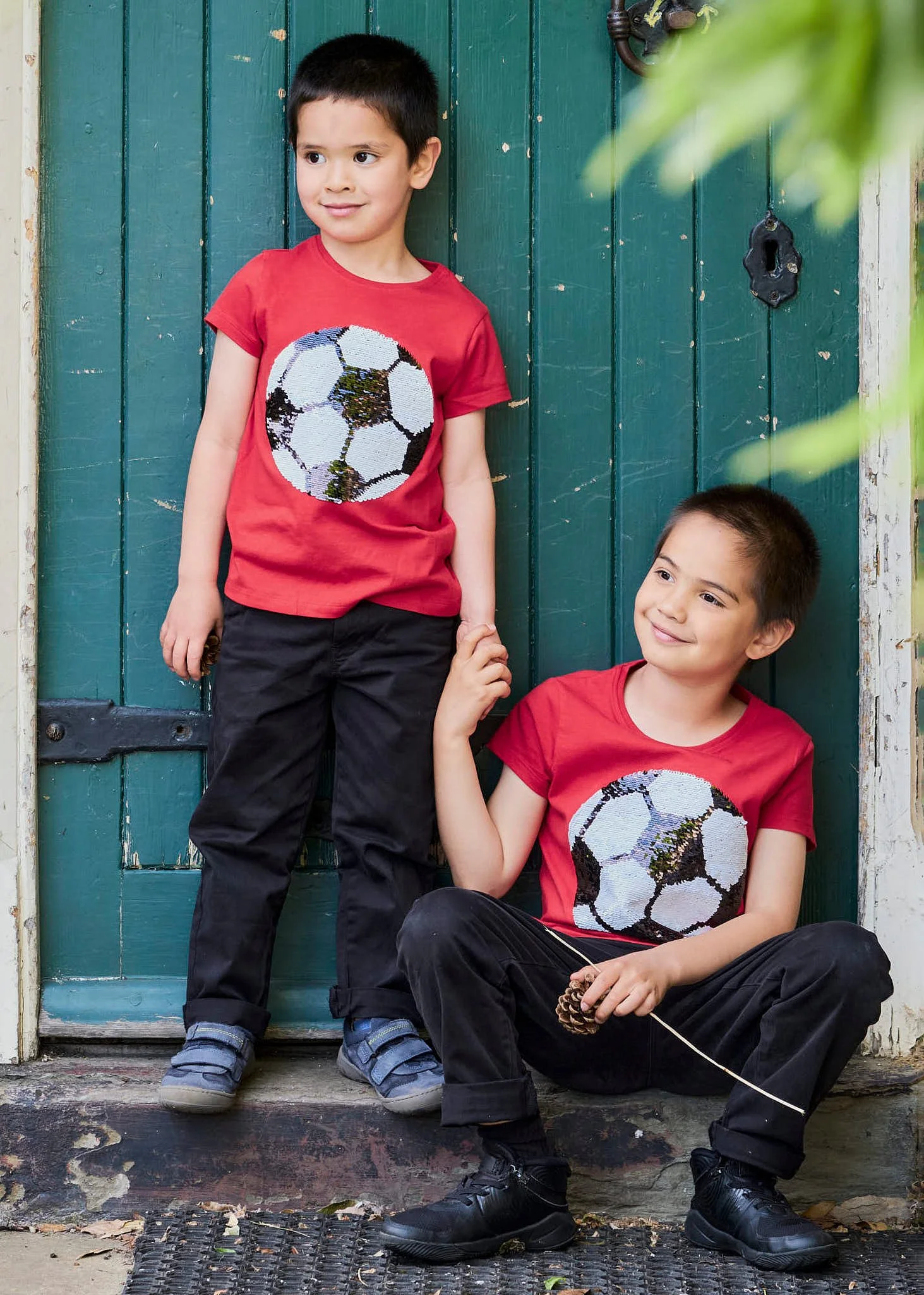 Two young boys wearing matching red T-shirts with a soccer ball design, black pants, and sneakers, posing in front of a turquoise wooden door. One boy is standing and the other is sitting, holding hands, with greenery in the foreground.