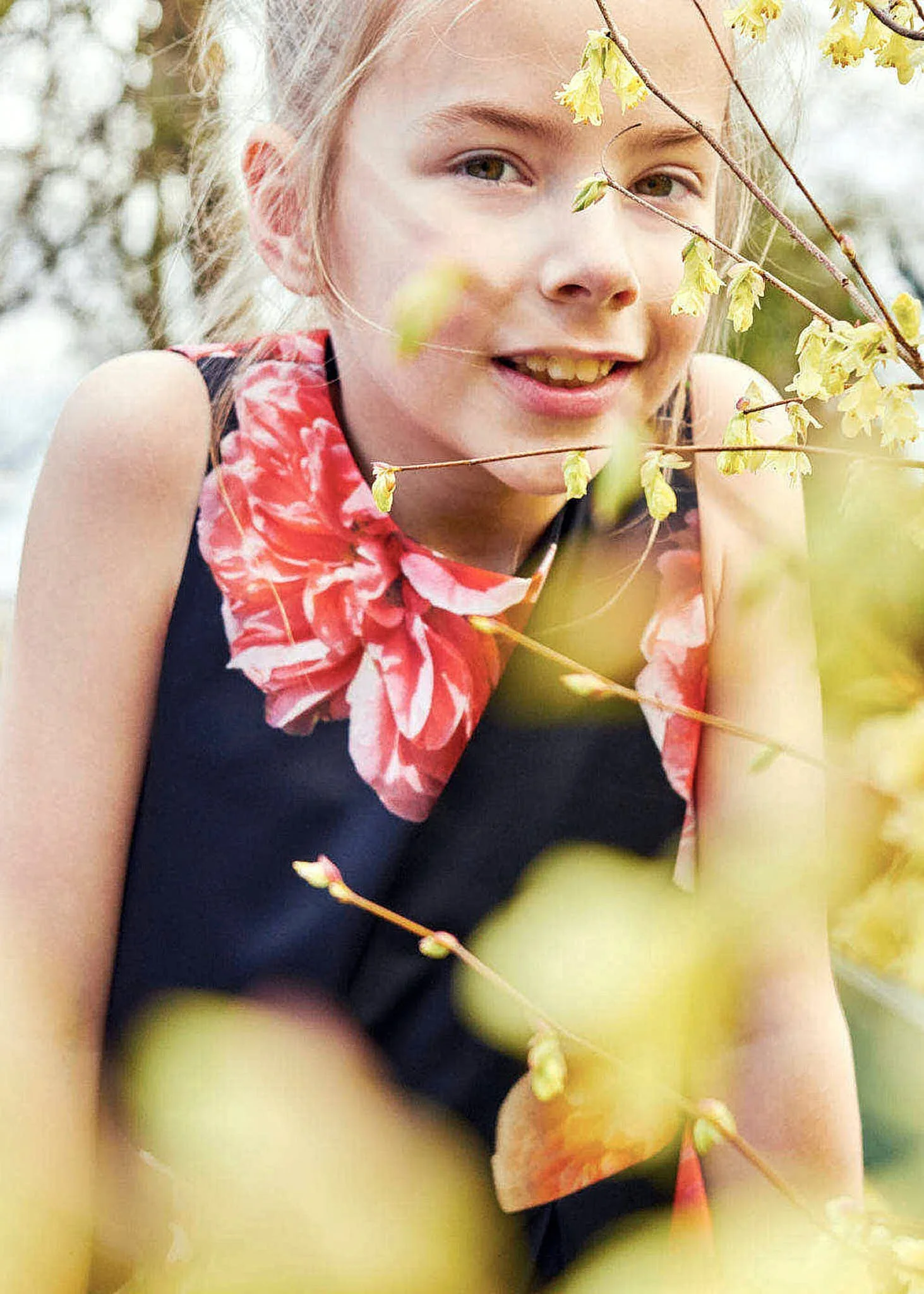 A young girl with blonde hair smiling, outdoors, surrounded by yellow flowering tree branches.