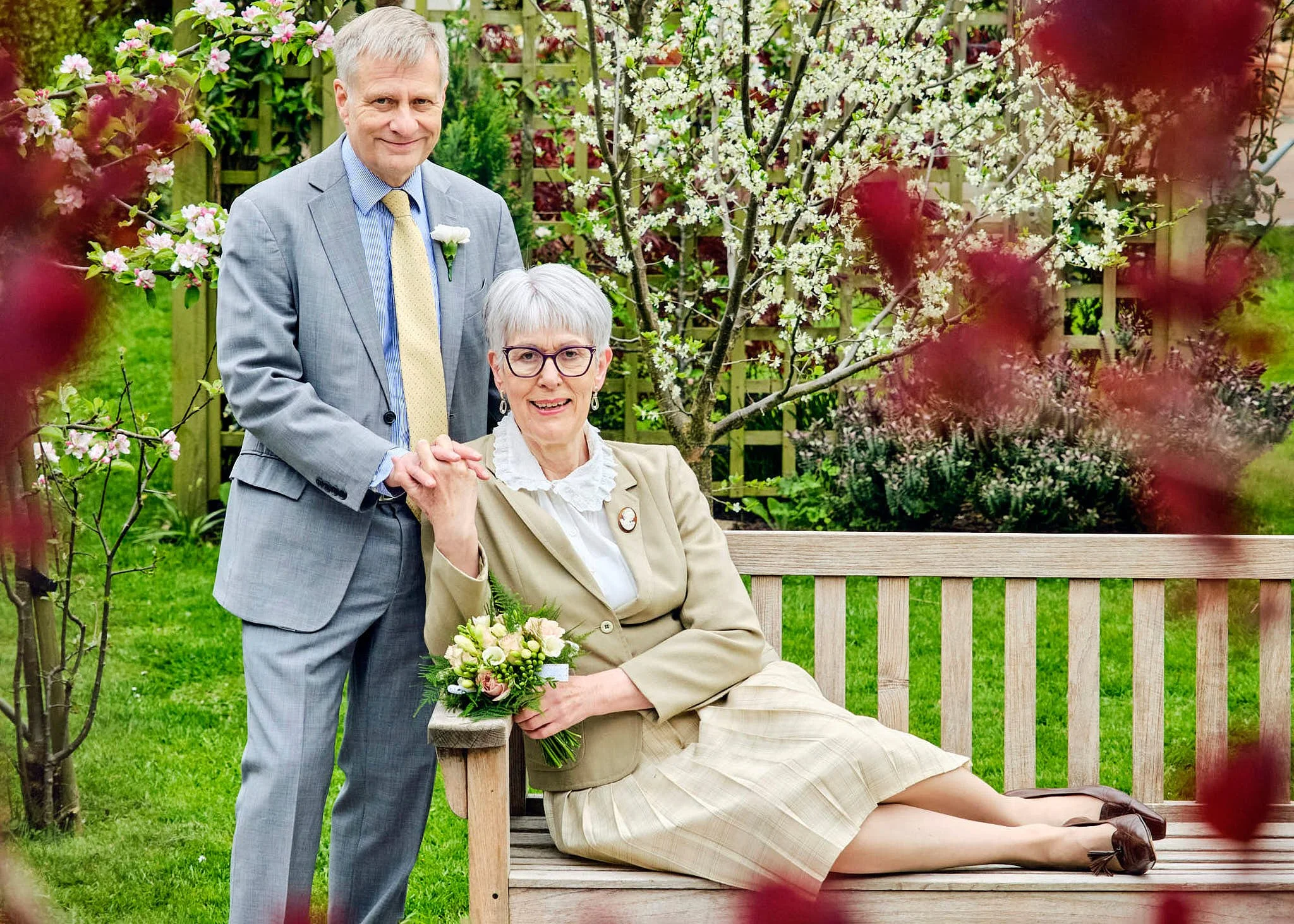 An elderly woman with short gray hair and glasses sitting on a wooden bench in a garden, holding a bouquet of flowers, while a middle-aged man in a gray suit with a yellow tie and white flower lapel pin stands behind her, holding her hand, both smiling.