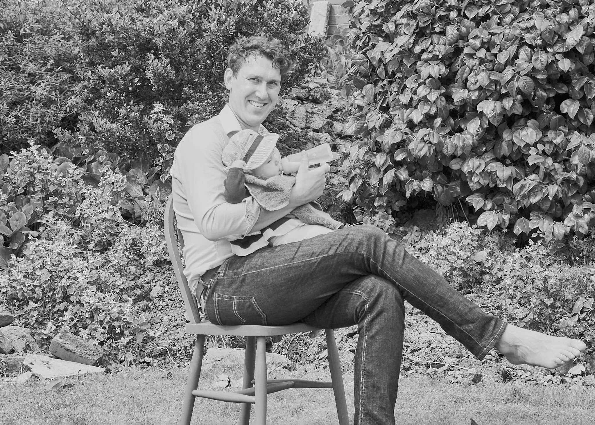 A man sits on a chair outdoors, holding a baby wrapped in a blanket. They are surrounded by lush bushes and plants.