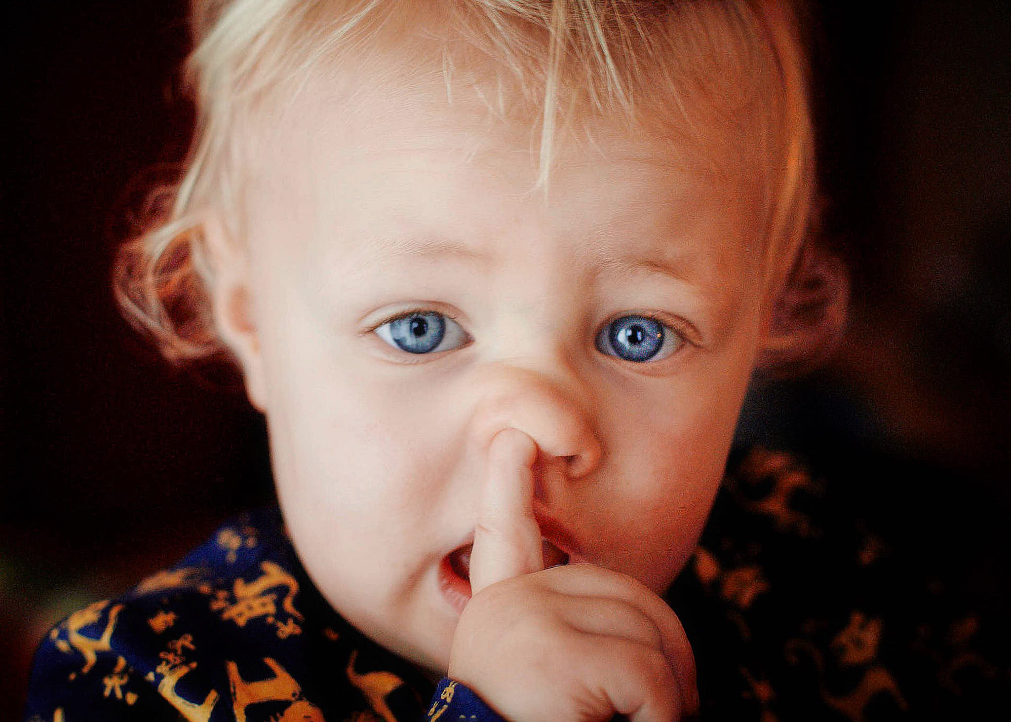 A young child with blue eyes, light blonde hair, and fair skin, holding their finger to their lips in a hush gesture.