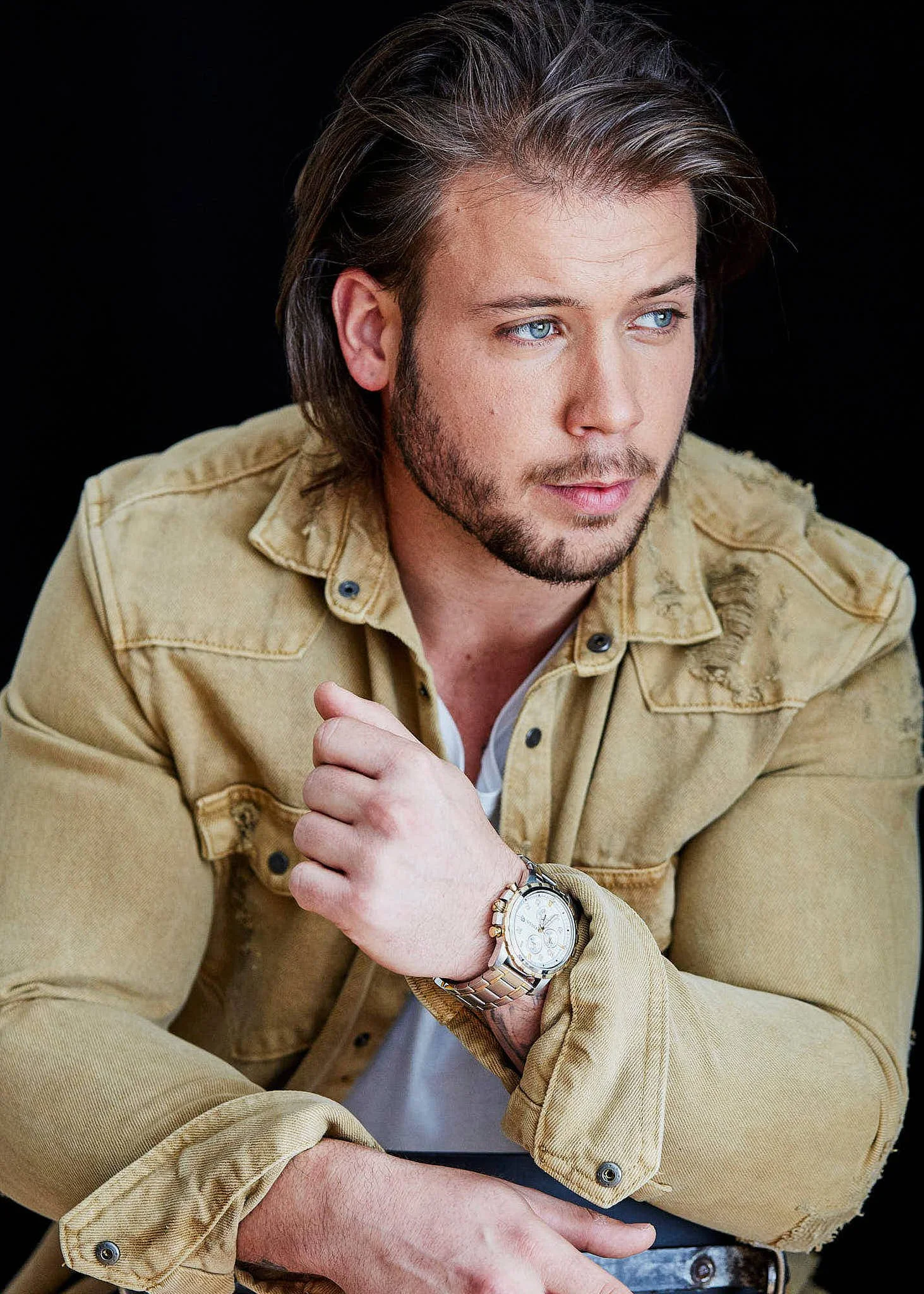 A man with blue eyes and brown hair, wearing a beige jacket and watch, sitting with arms crossed, looking to the side against a black background.