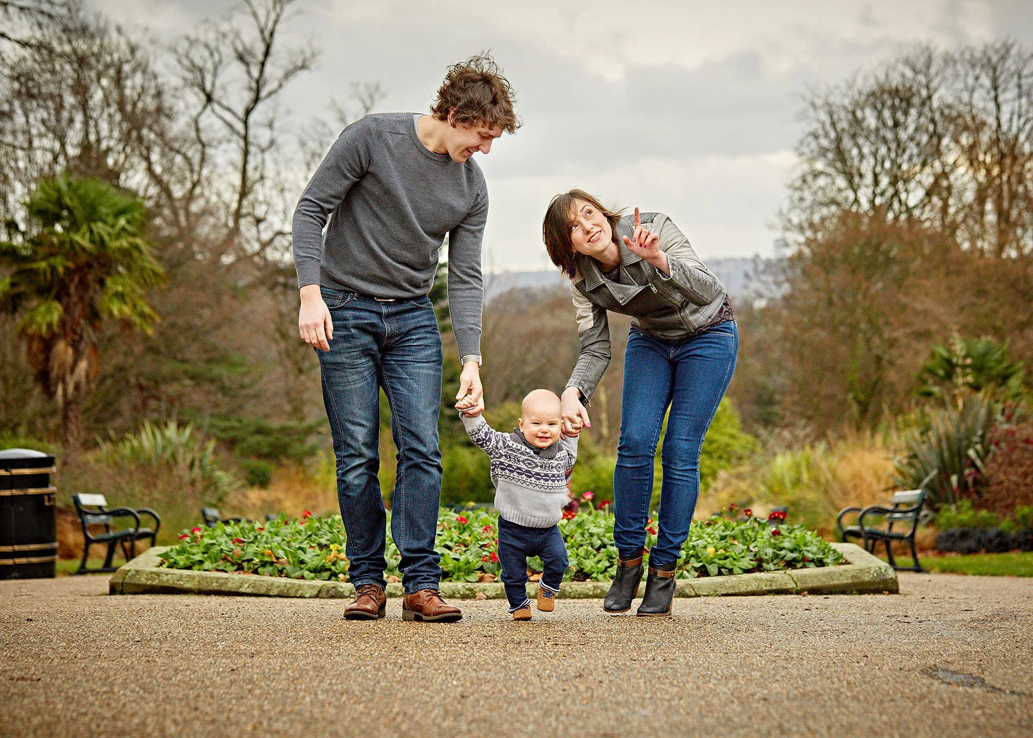 Two women and a young girl are walking in a garden, holding hands, with trees and flower beds in the background.