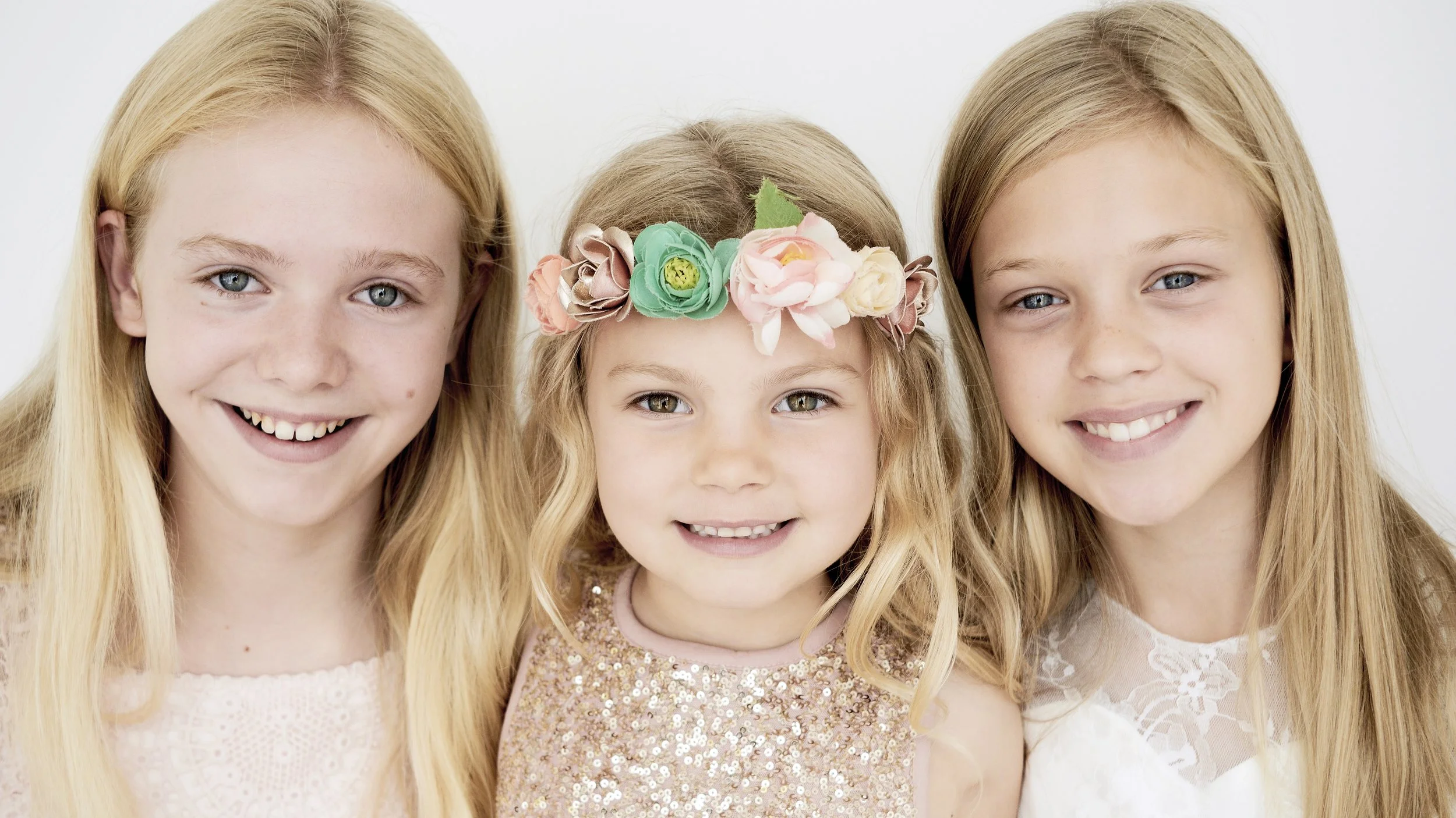 Three young girls with blonde hair and blue eyes smiling, with the girl in the middle wearing a floral headband.