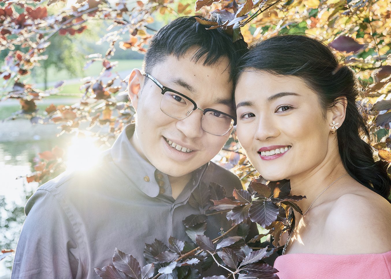 A smiling Asian couple posing outdoors near a body of water, with fall foliage in the background. The woman has dark hair and is wearing a pink off-the-shoulder top and floral earrings. The man is wearing glasses and a button-up shirt. They are close together, with sunlight shining behind them.
