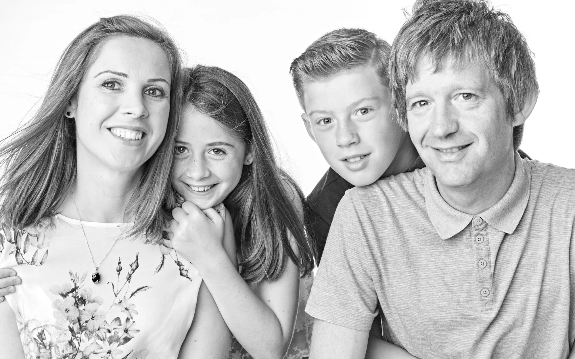 A black and white photo of a smiling family group of four, including a woman, a girl, a boy, and a man, posing closely together against a plain backdrop.