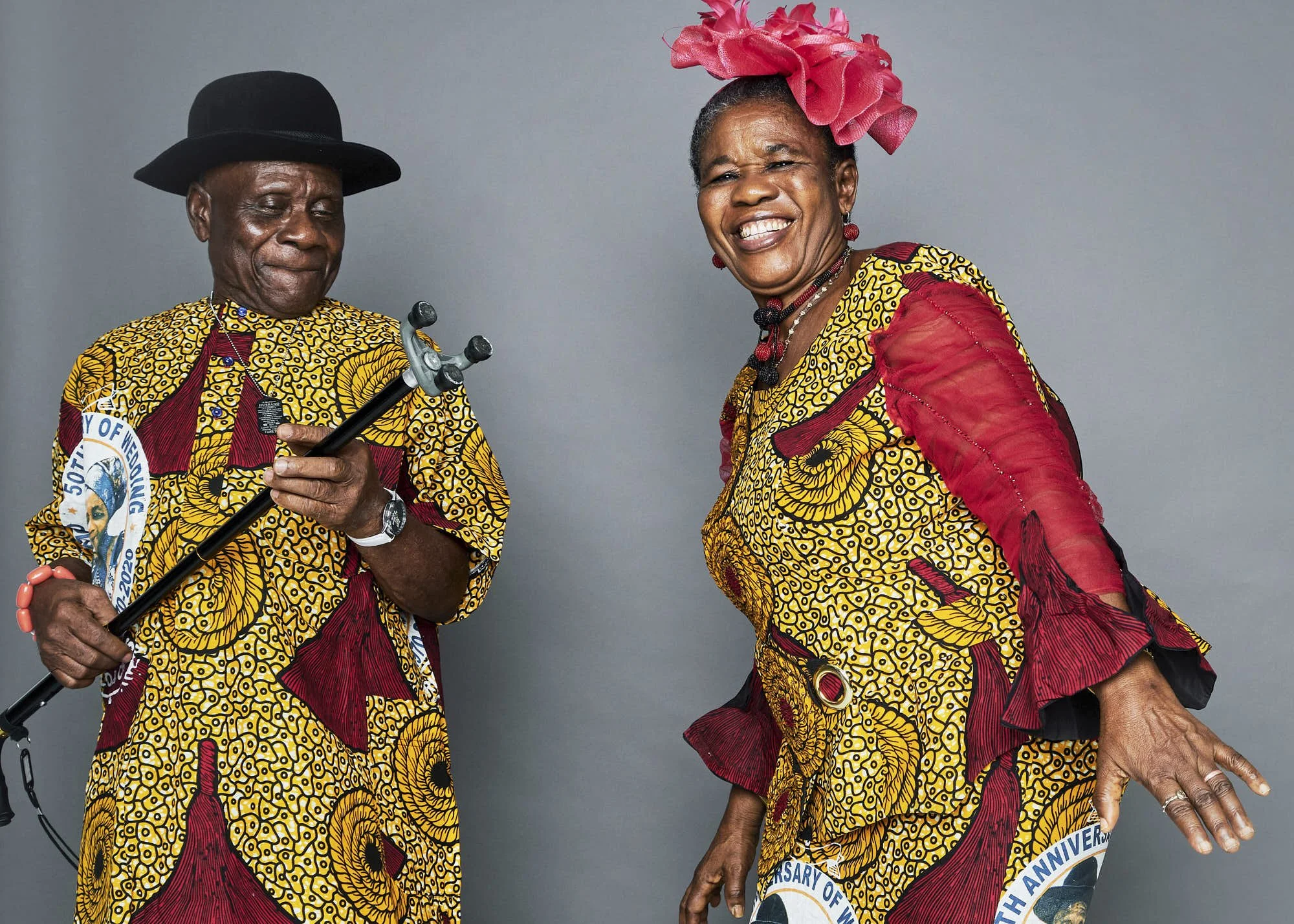 An elderly man and woman dressed in colorful, patterned African attire, celebrating, with the man holding a cane and the woman smiling.