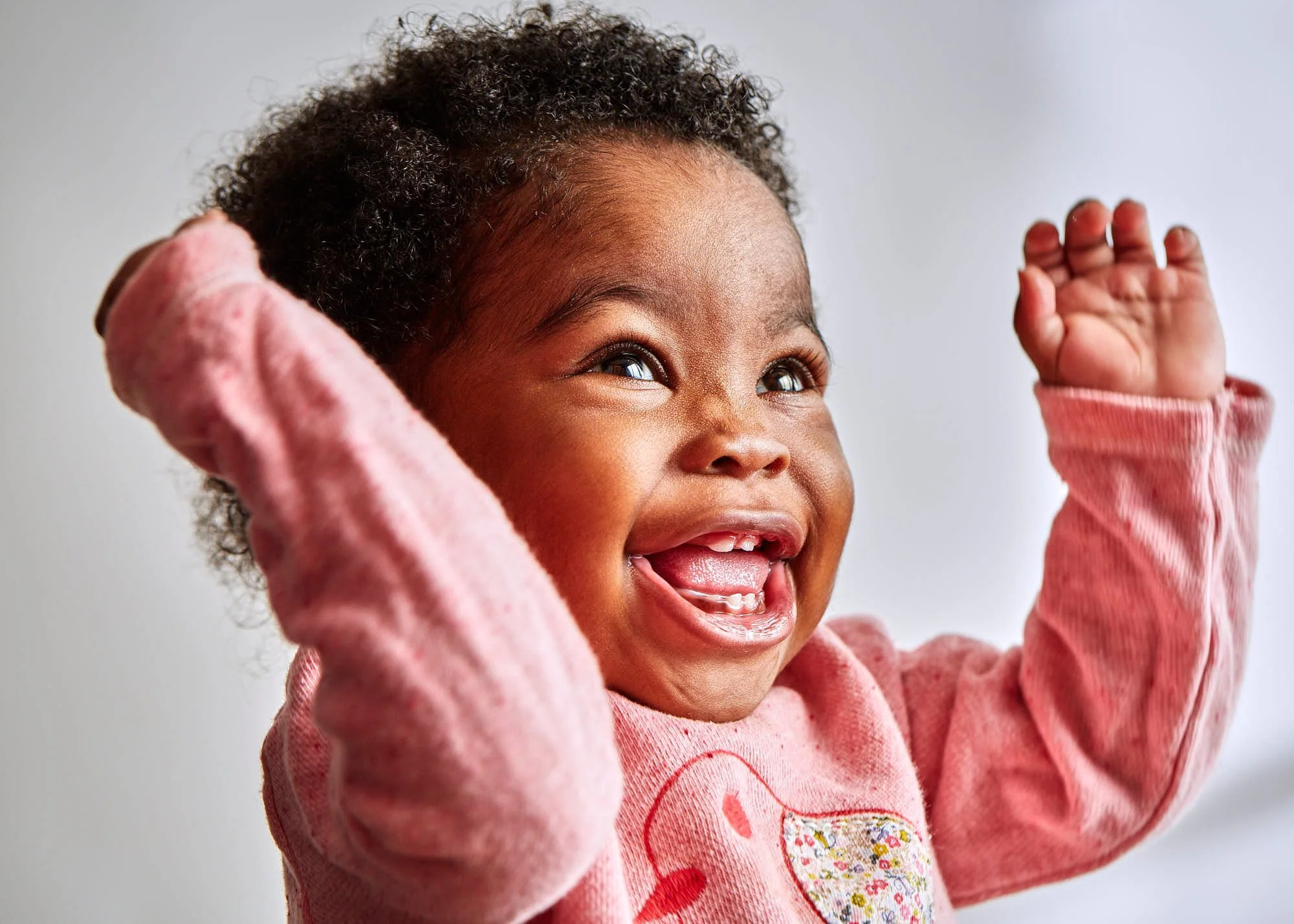 A young girl with curly hair smiling and raising her hand, wearing a pink sweatshirt.