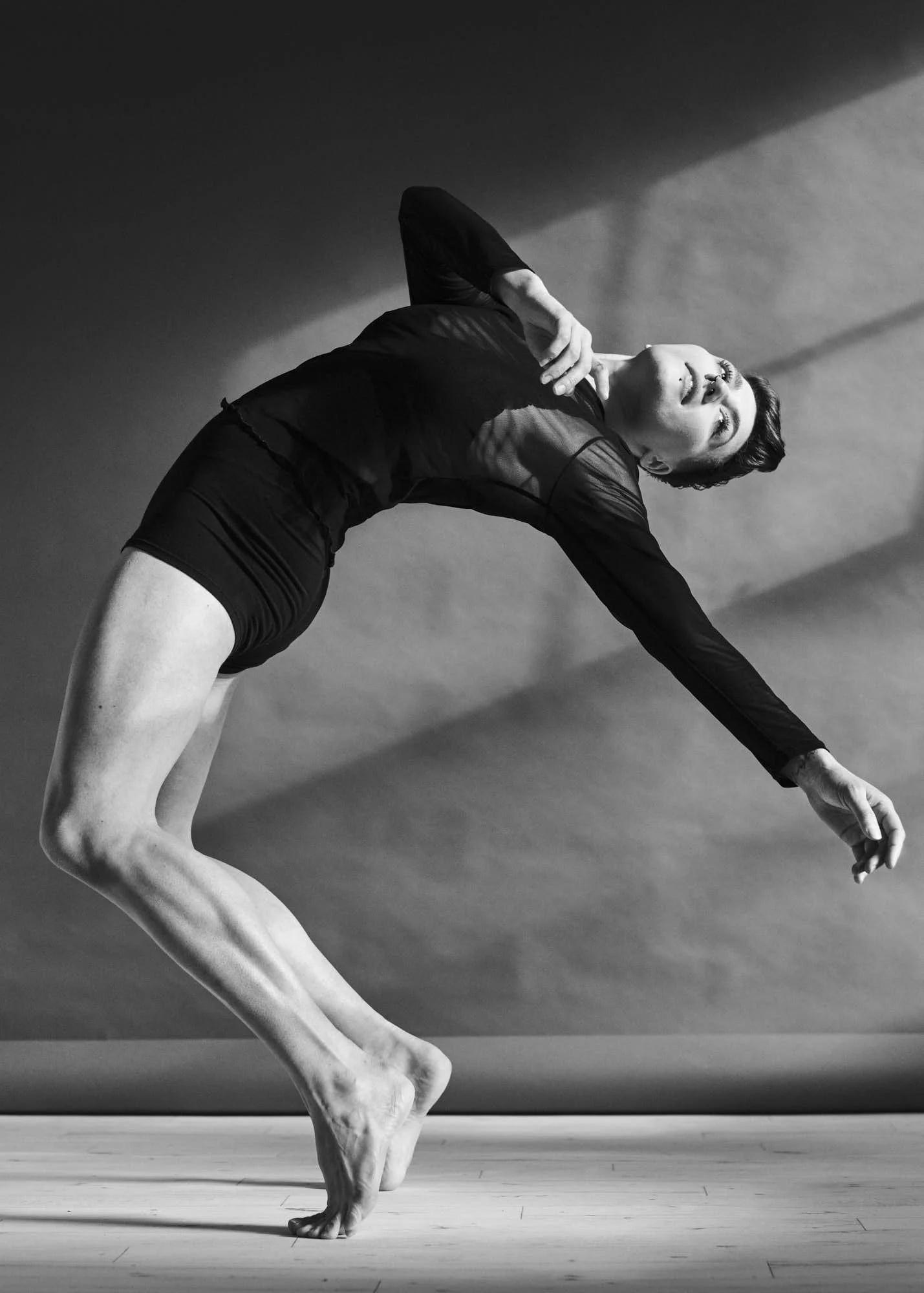A person performing a handstand in a dance or yoga pose, with one leg bent and slightly raised, wearing a black long sleeve shirt and shorts, in a studio with wooden floor and plain background.