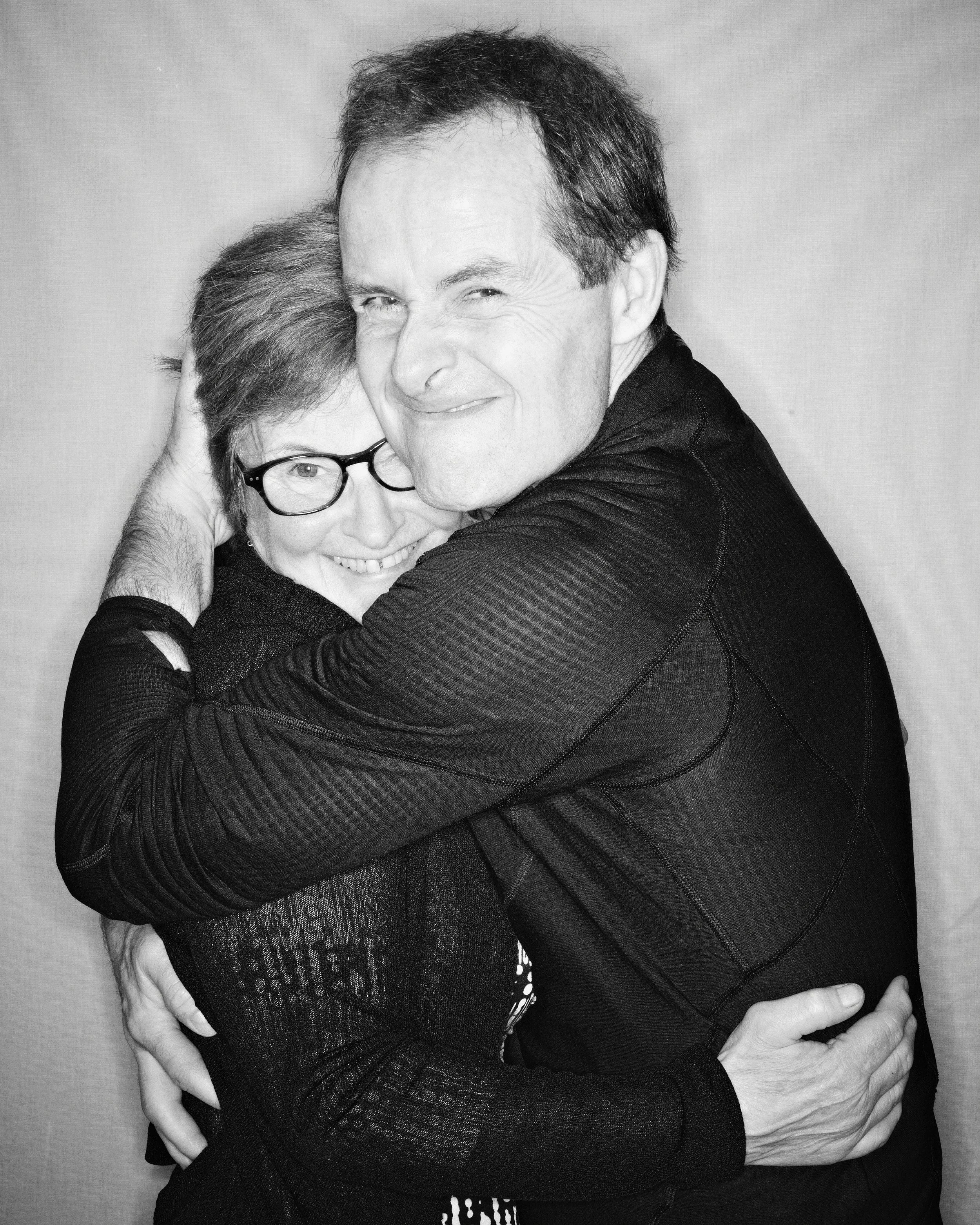 A black and white photograph of a man and a woman hugging. The man has dark hair and is smiling with his eyes closed, while the woman, wearing glasses, is also smiling. They are embracing each other tightly.