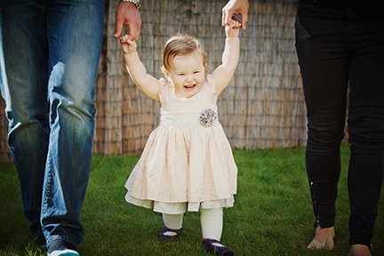 A young girl in a cream dress holding hands with two adults in a backyard with a wooden fence and green grass.