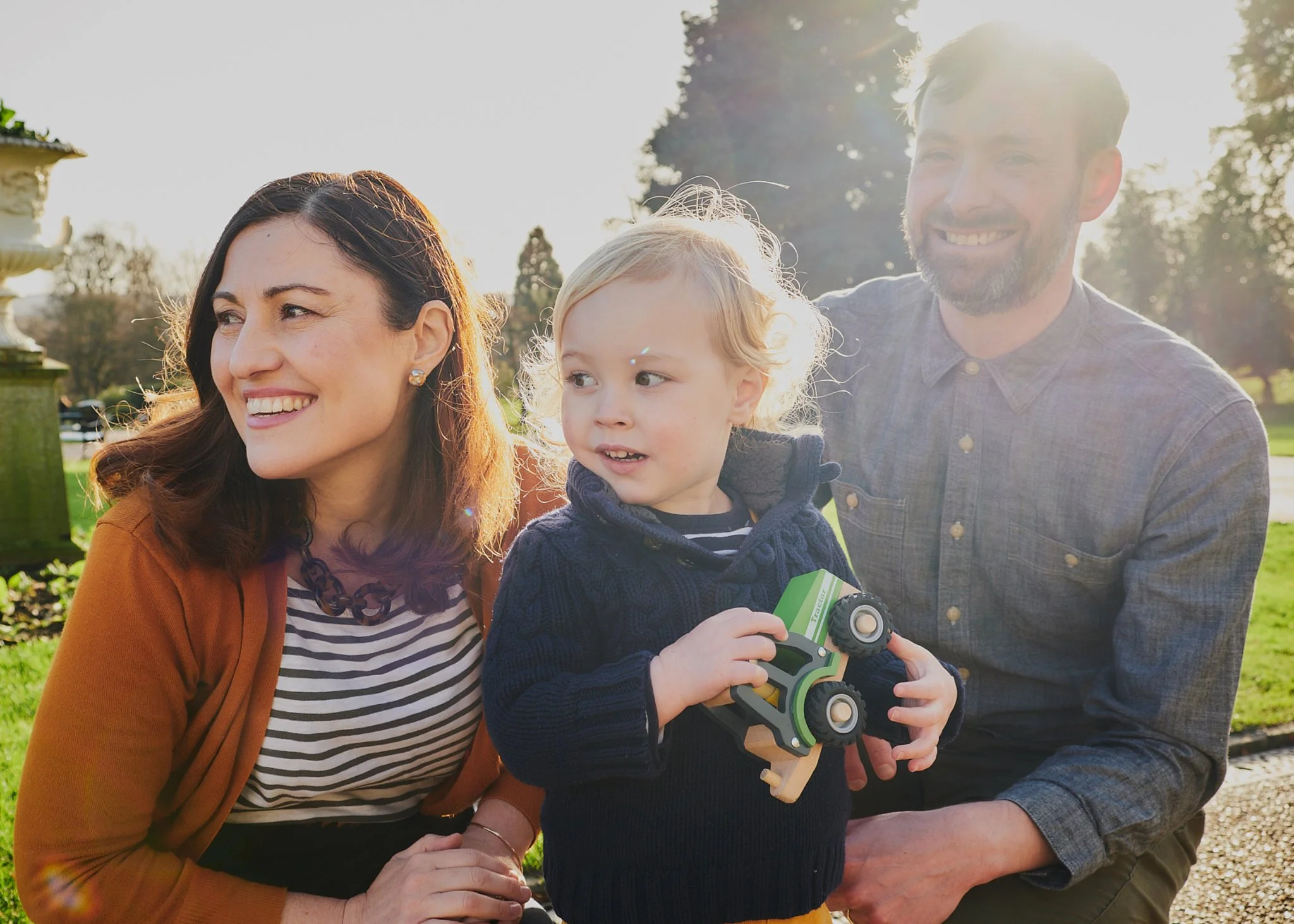 A family of three smiling outside in a park during the day. The woman has brown hair, the man has a beard, and the little boy has blonde hair and is holding a toy vehicle.