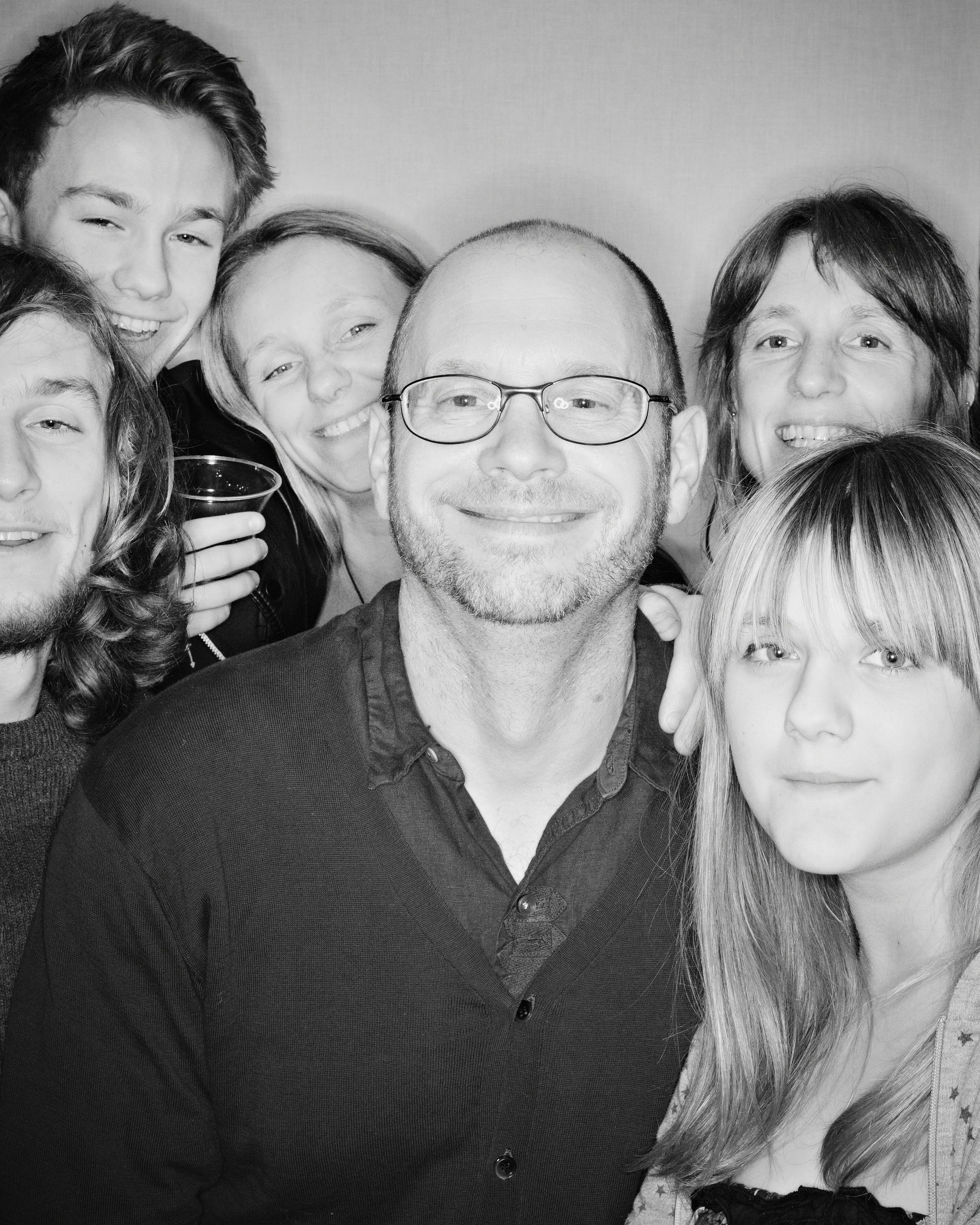 Black and white photo of a group of people closely gathered, smiling and looking at the camera. The group includes both men and women of various ages.