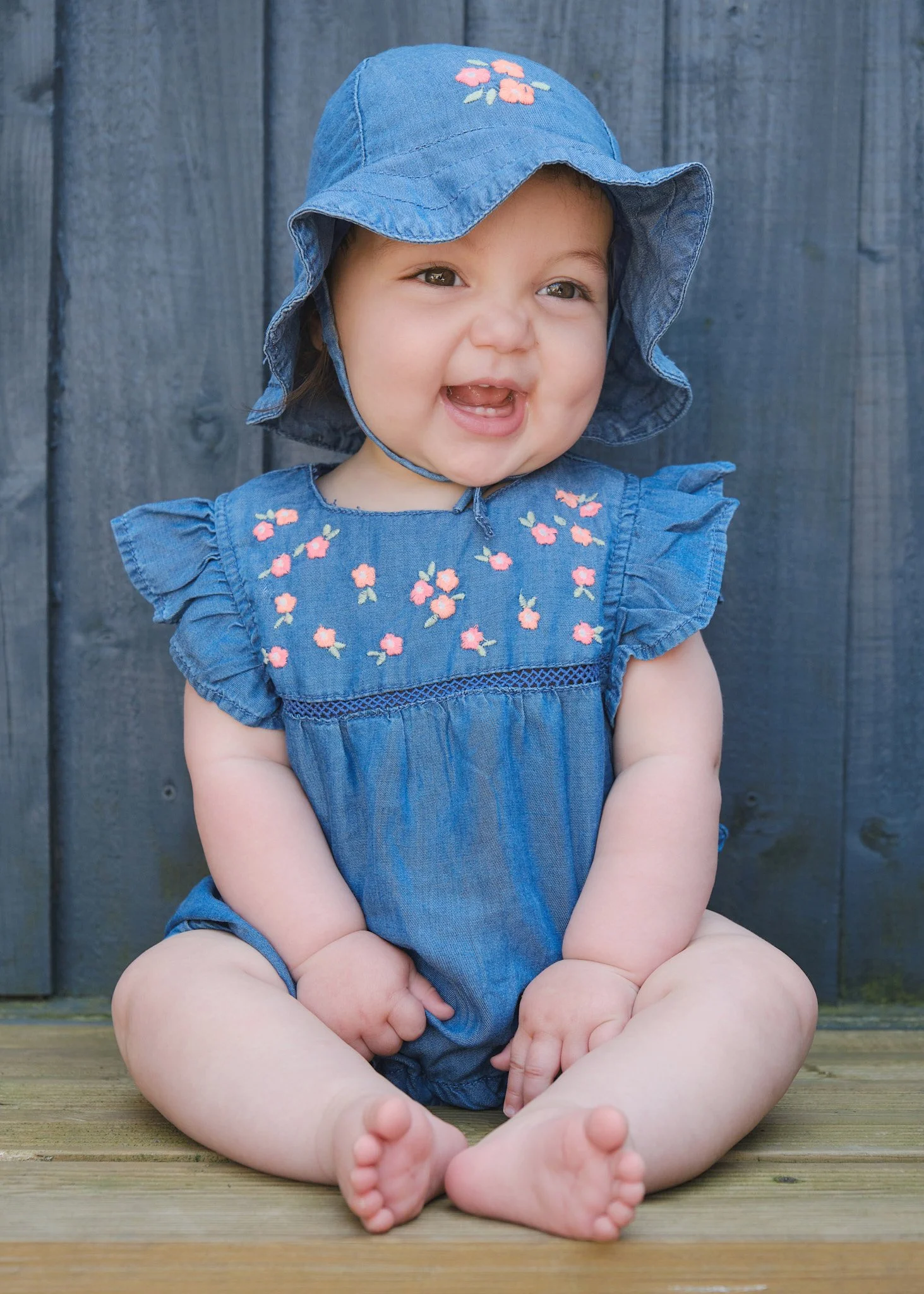 A young girl sitting outdoors on a wooden surface, wearing a denim dress and a matching denim sun hat with pink embroidery, smiling with her mouth open.