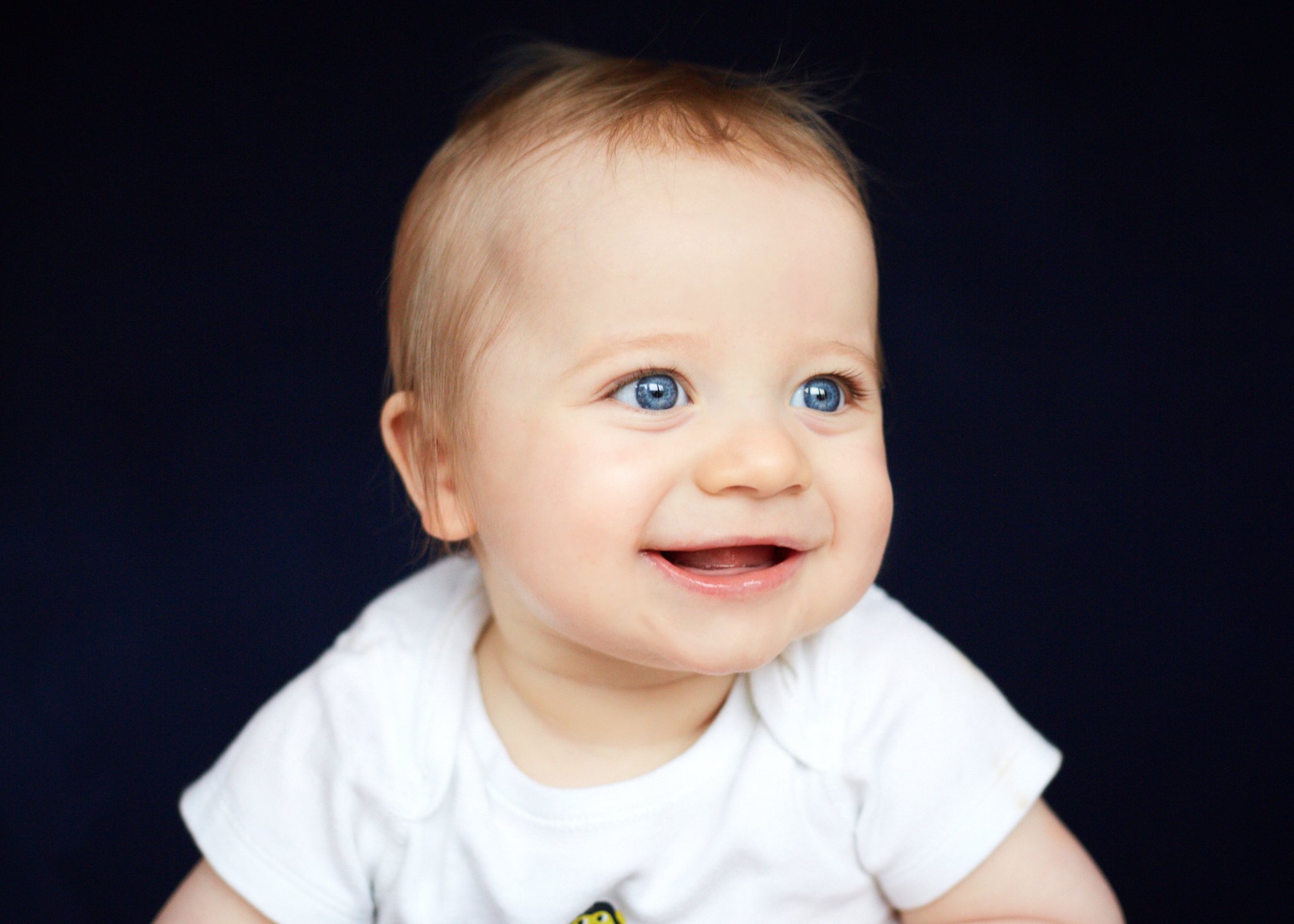Close-up of a smiling baby with blue eyes and light brown hair, wearing a white shirt, against a dark background.