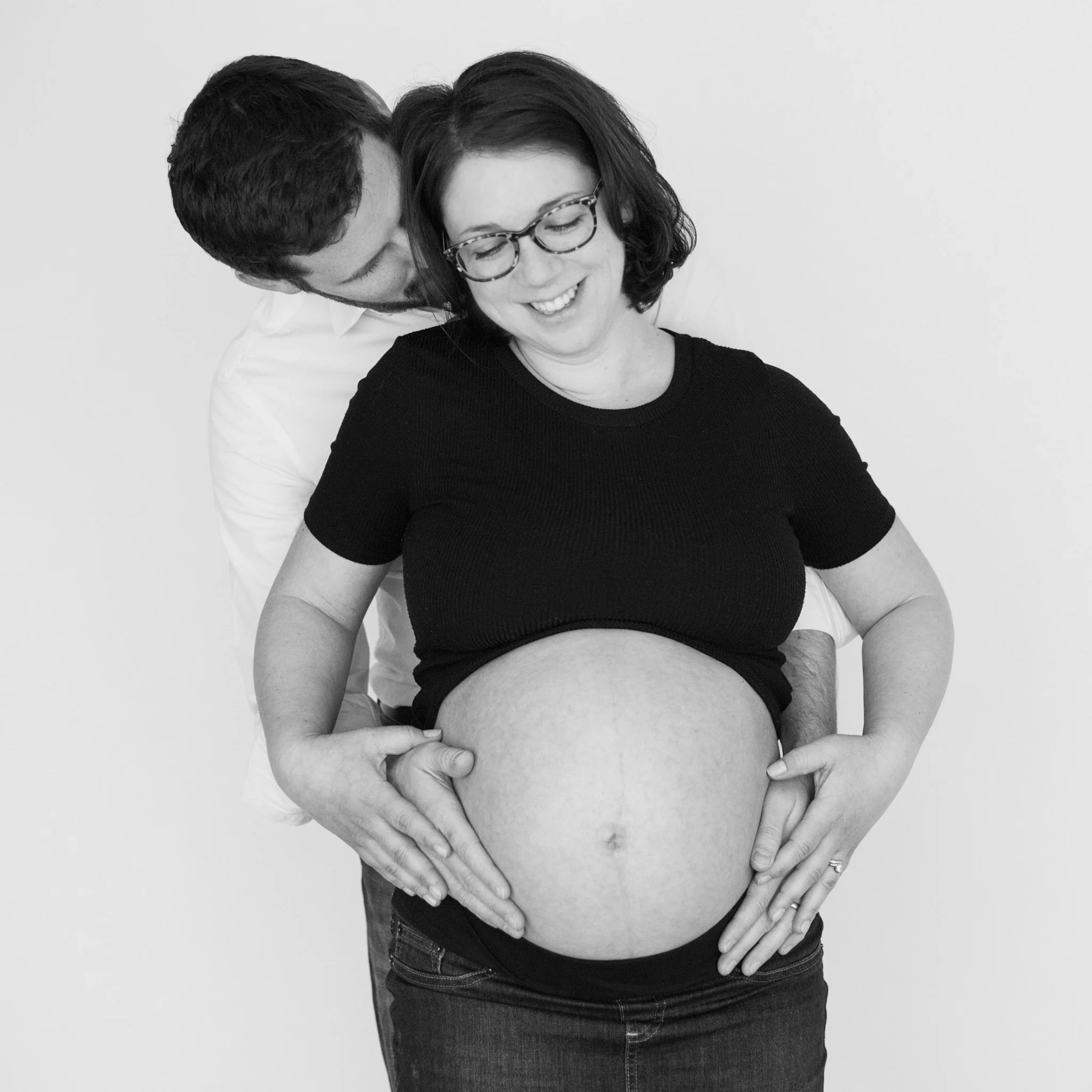 A man and a woman share an intimate moment, with the man whispering into the woman's ear from behind. The woman is smiling and wearing glasses, while the man has his face close to her ear, eyes closed. The woman's hands are on her pregnant belly.