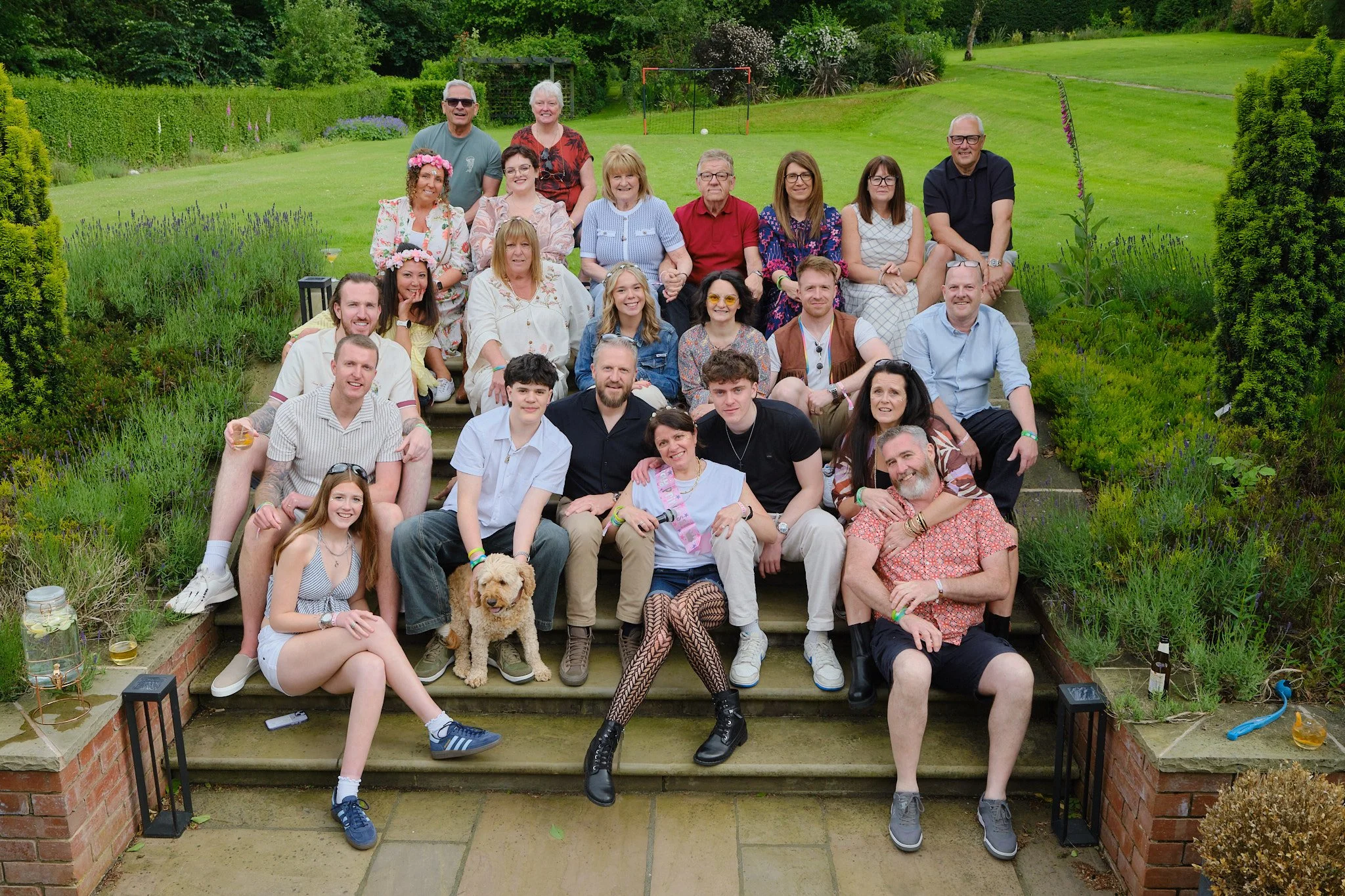 Group of 19 people sitting and standing on outdoor stone steps in a garden, with some holding drinks, and a dog in front, during daytime.