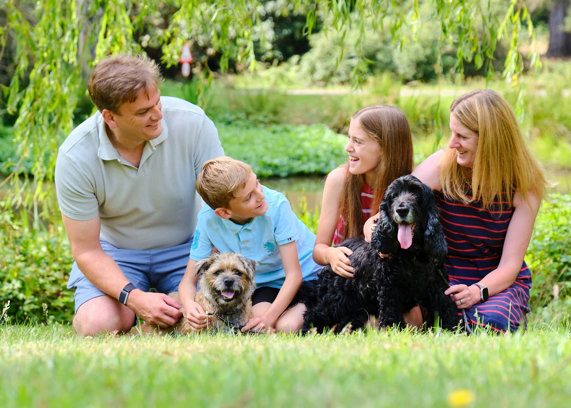 A smiling family of five with two dogs sitting outdoors in a lush, green park with trees and a small pond, enjoying a sunny day.