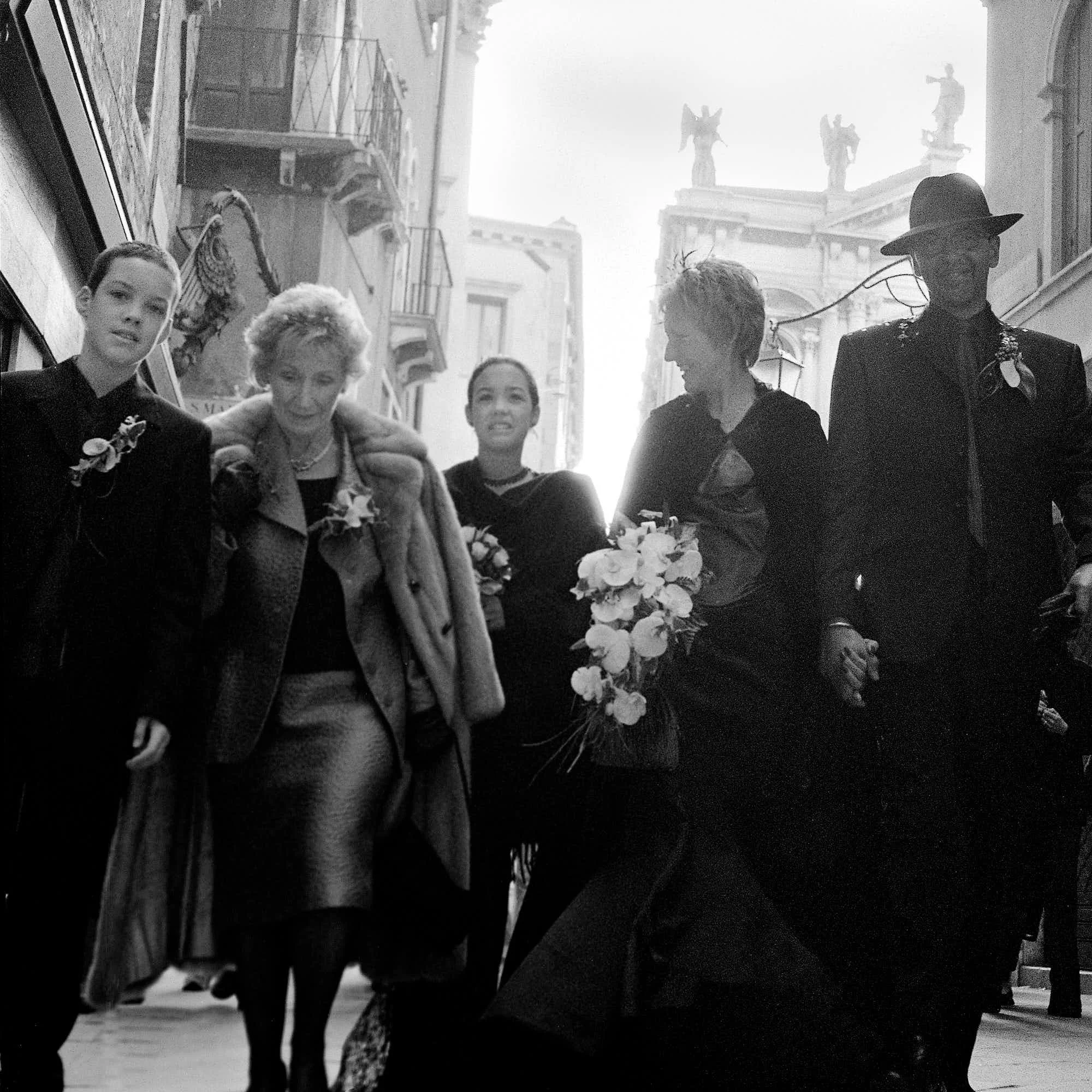 A black and white photograph of five people walking down a city street with ornate buildings and statues on the rooftops in the background. The group includes two women and three men, with one man wearing a wide-brimmed hat and sunglasses, and some o