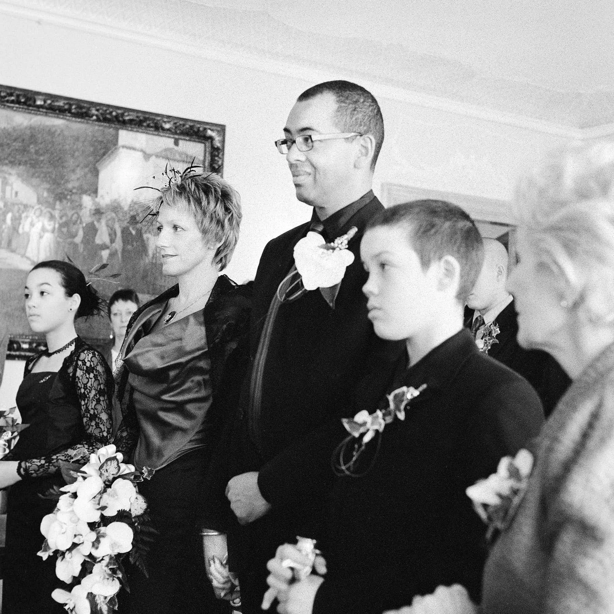A black and white photo of a man in a suit and tie, wearing glasses, standing in a line among several young girls, with a woman beside him, all dressed formally during an indoor event with a painting on the wall in the background.
